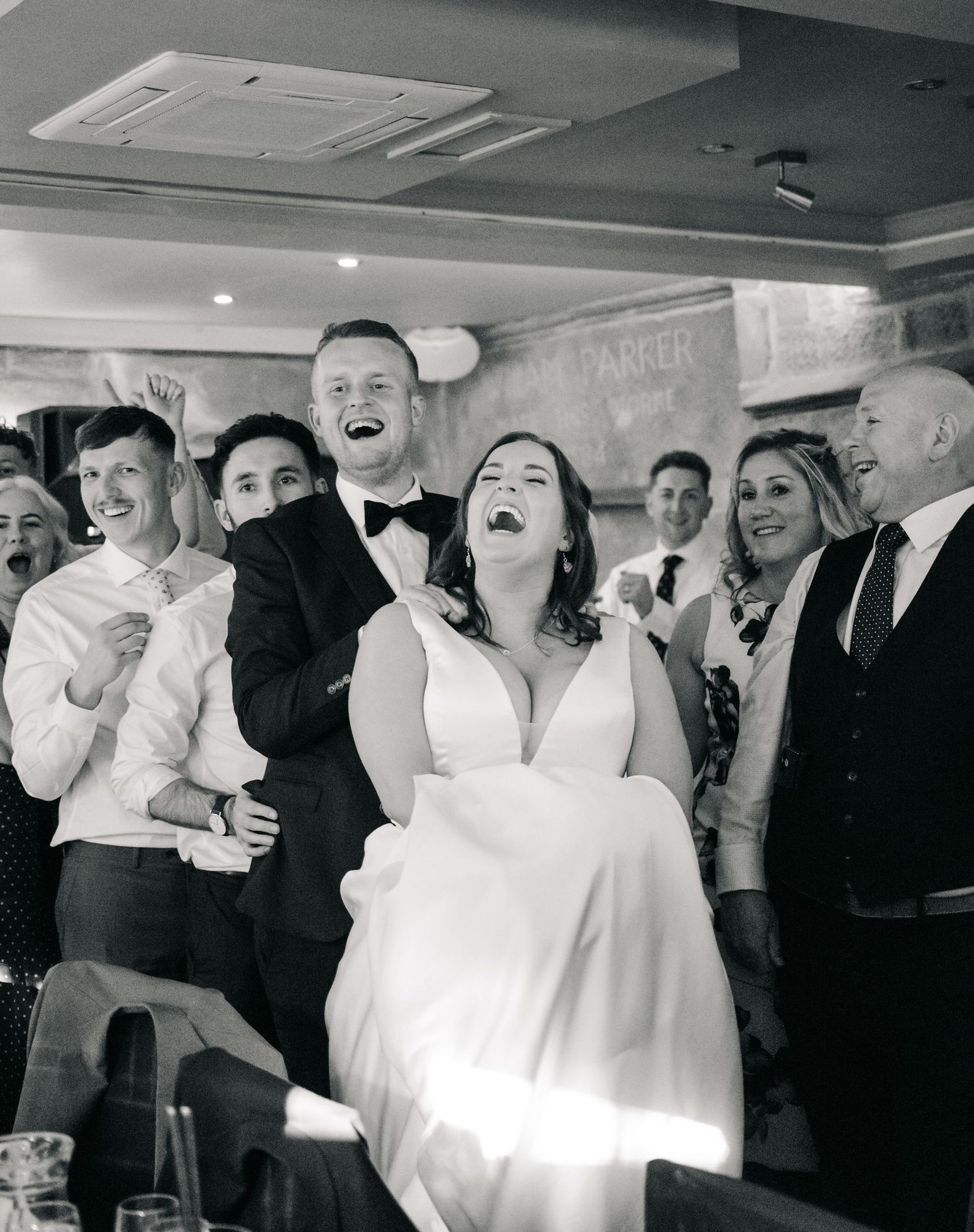 Black and white photo of laughing bride in wedding dress surrounded by joyful guests at a wedding reception.