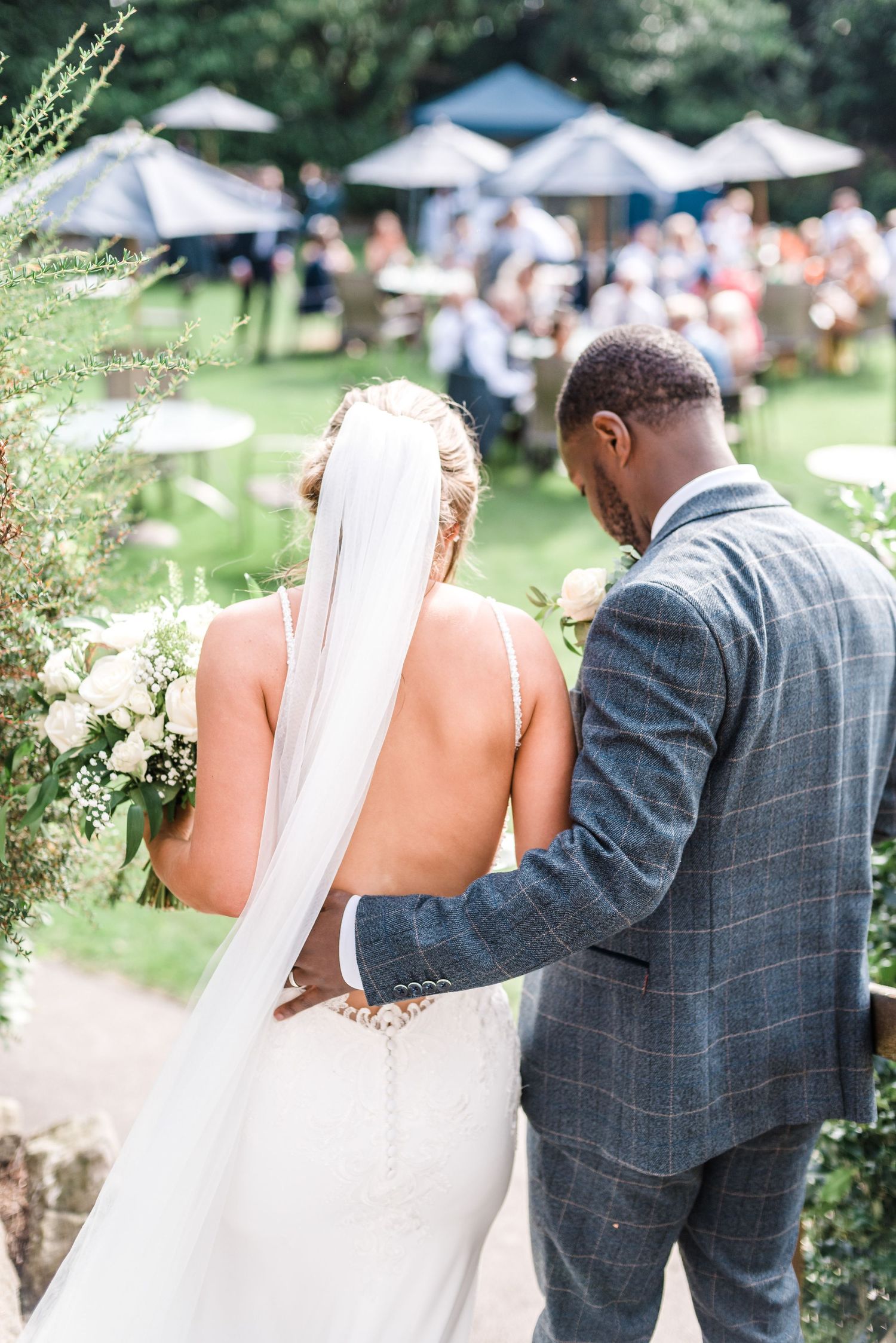 A couple shares an intimate moment in a garden setting with white tents in the background.