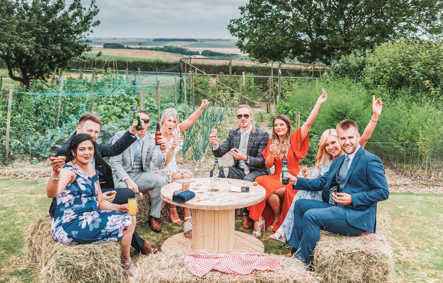 Wedding guests celebrating on hay bales in colorful garden setting.