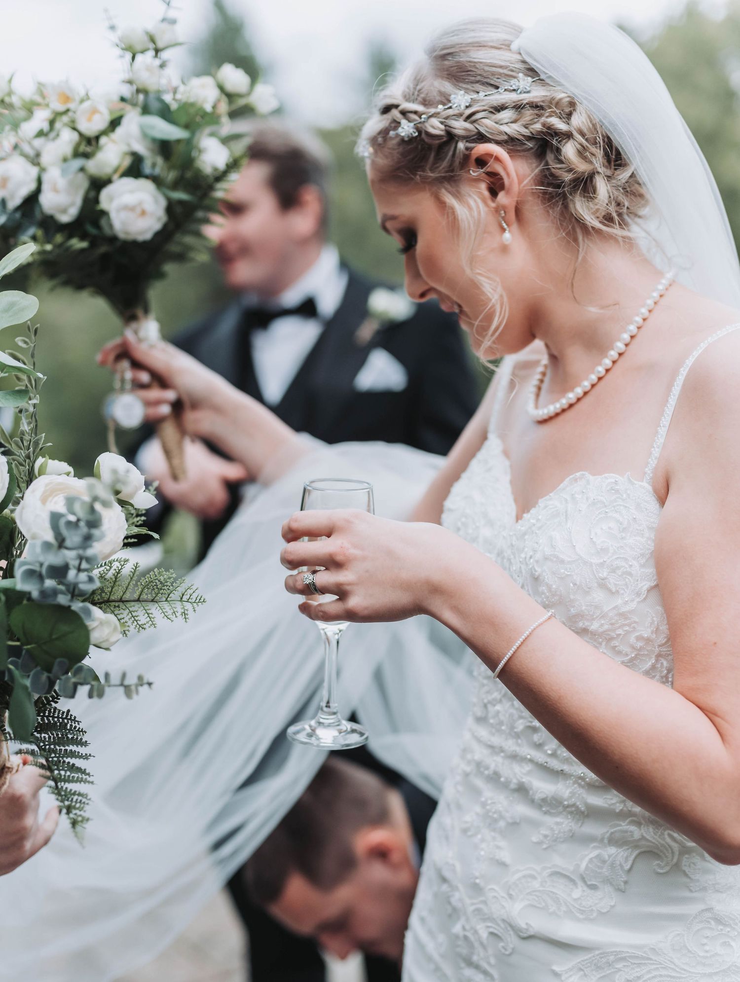 Bride in lace dress with braided hair holds champagne glass near floral arrangement.