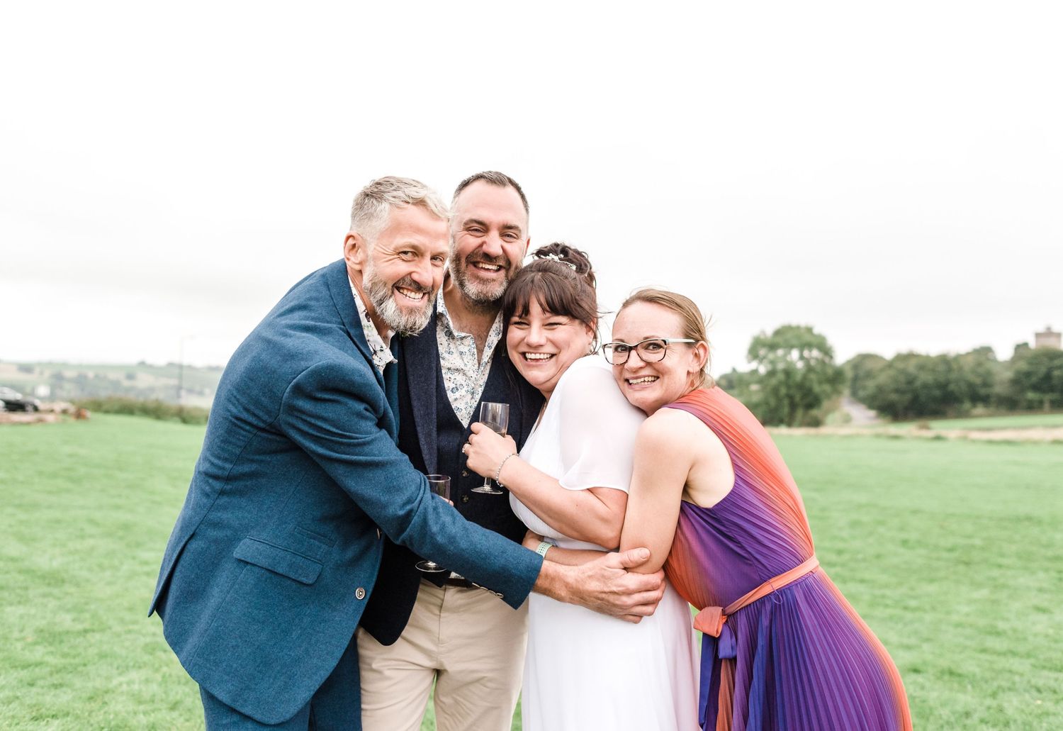 Wedding party embracing and laughing together on grassy field.