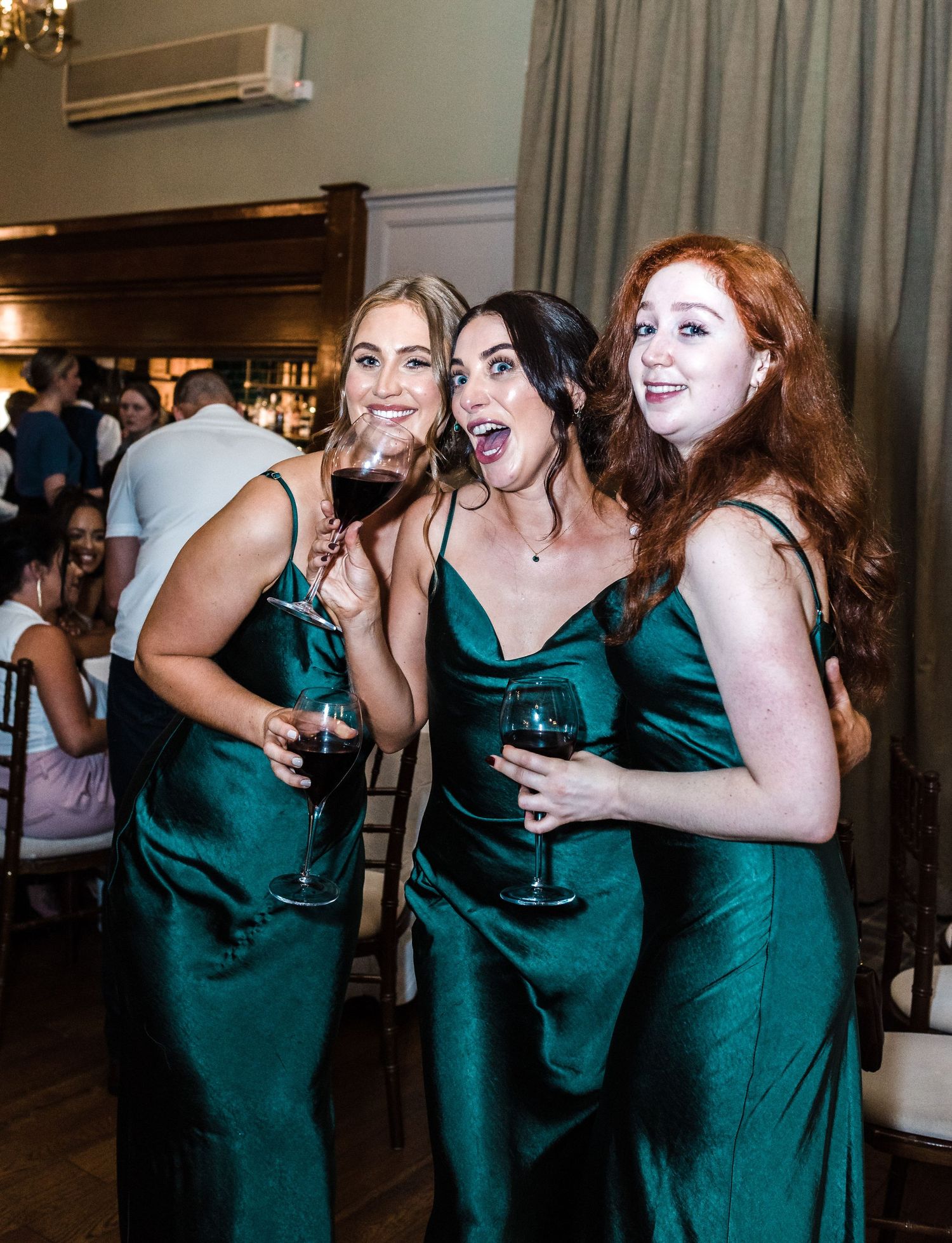 Three bridesmaids in emerald green satin dresses sharing laughs during wedding reception.