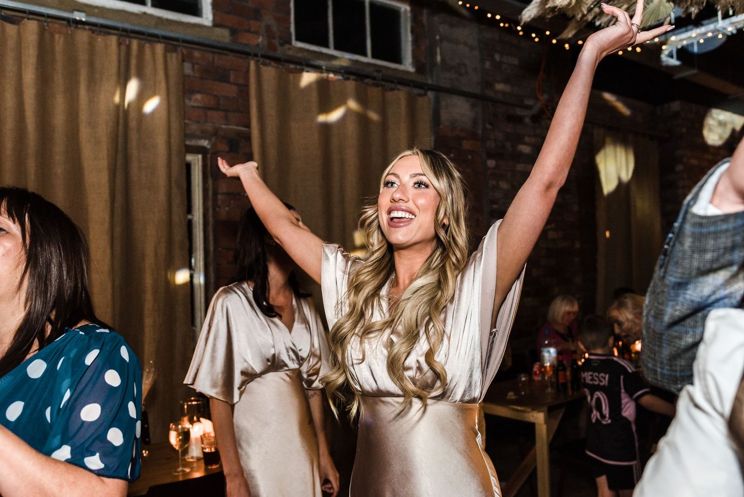 Woman in champagne silk dress dancing with raised arms at wedding reception.