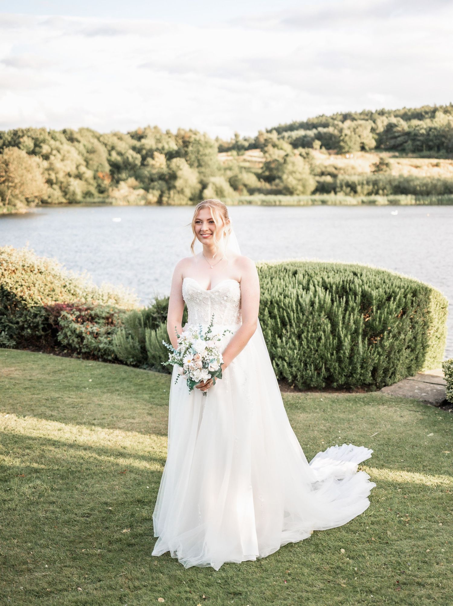 Bride in flowing white gown poses with bouquet by lakeside on manicured lawn.