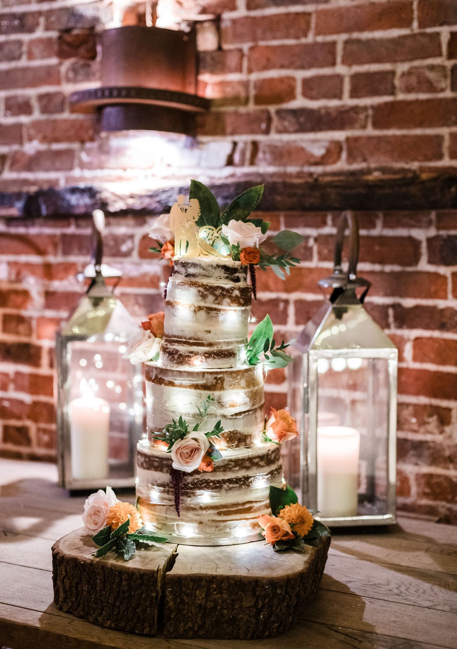A rustic semi-naked wedding cake decorated with fresh flowers sits on a wood slice against an exposed brick wall.