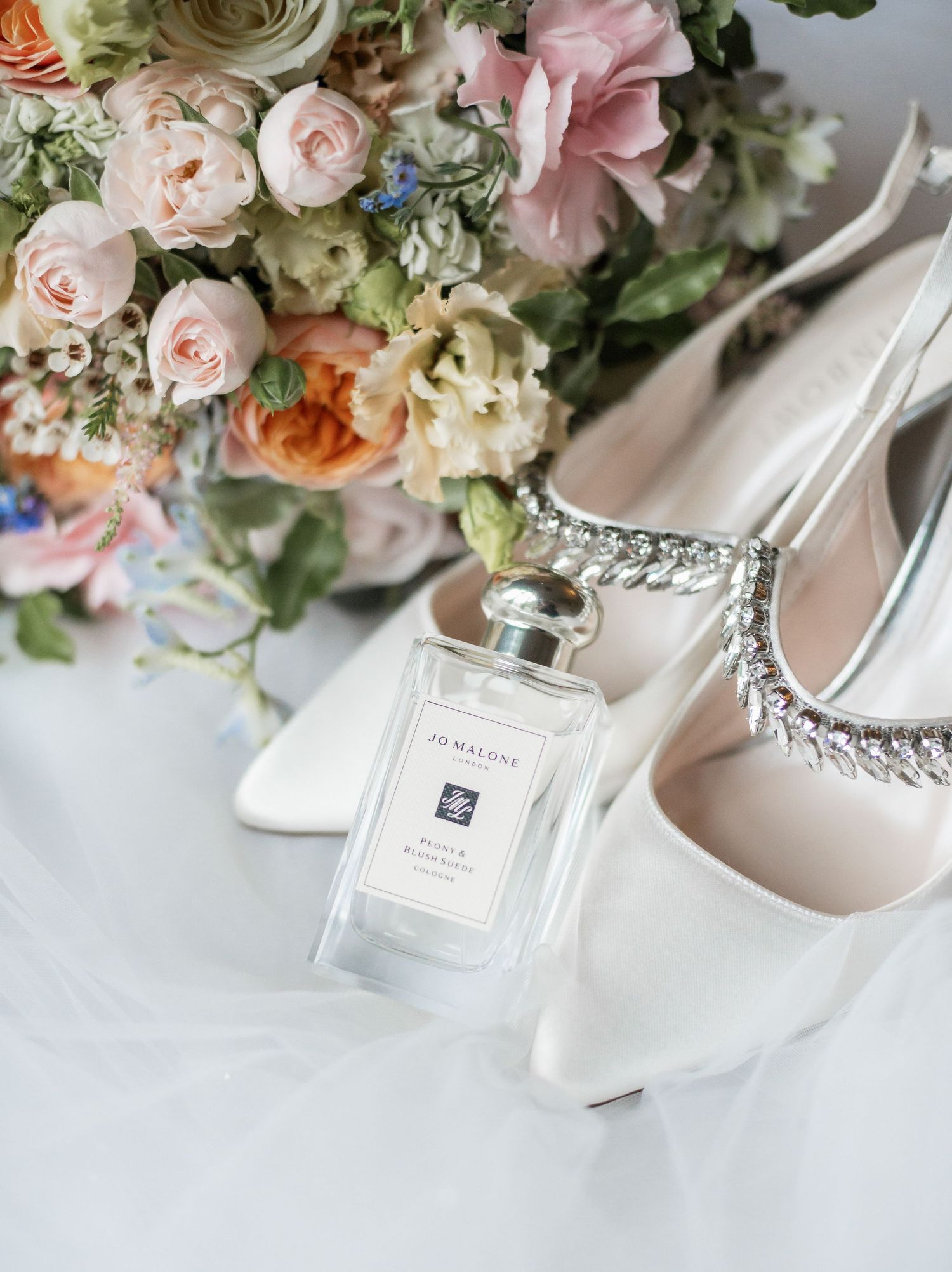 Wedding details featuring bridal shoes, perfume bottle and pastel floral bouquet on white background.