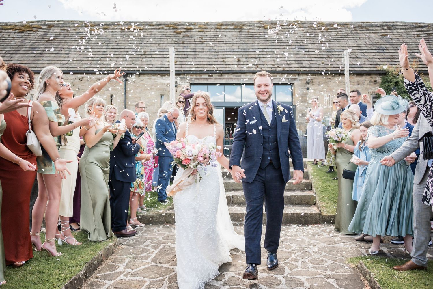Wedding guests throw confetti as newlyweds exit their ceremony on a stone pathway.