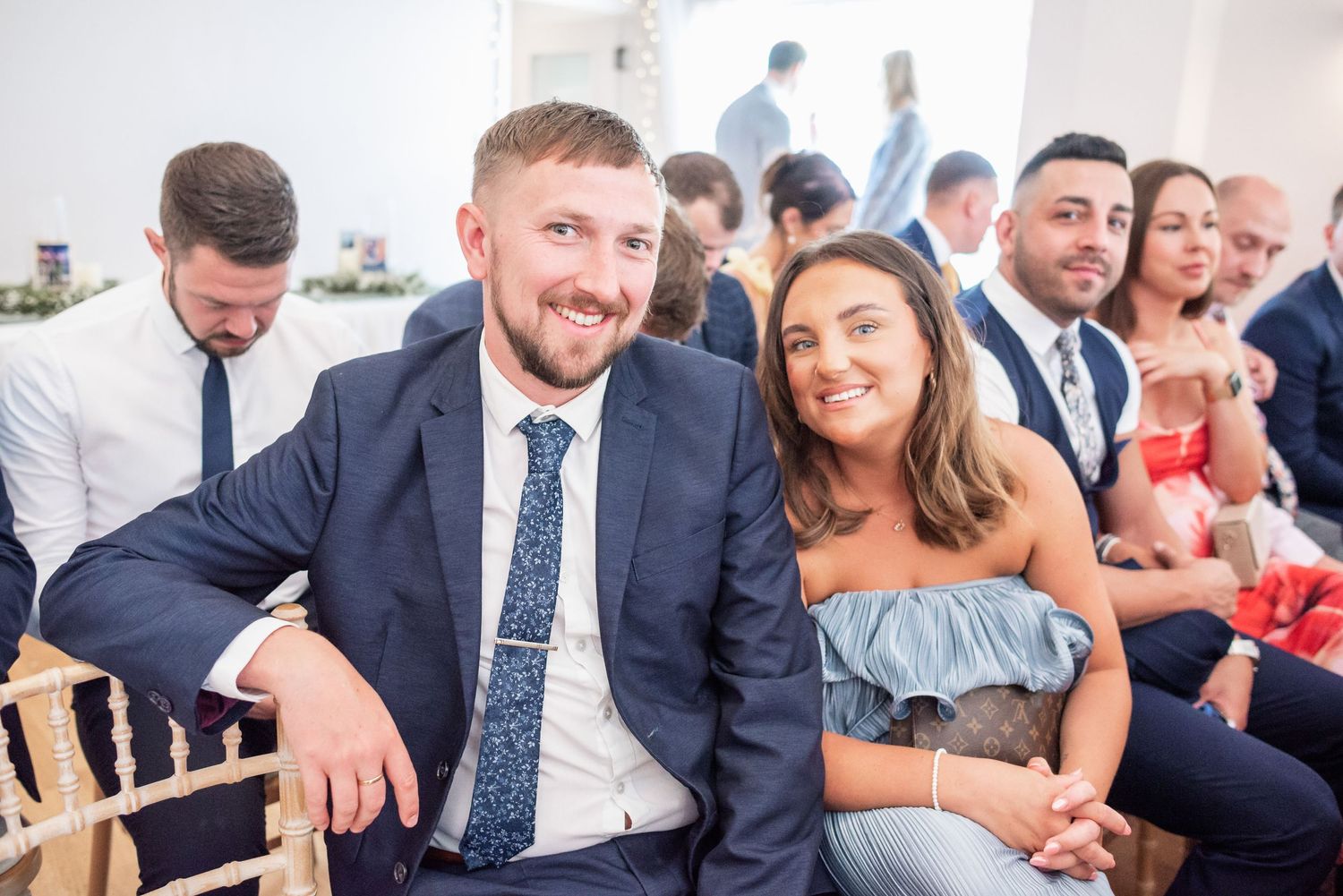 Wedding guests smile while seated in wooden chairs at an indoor venue.