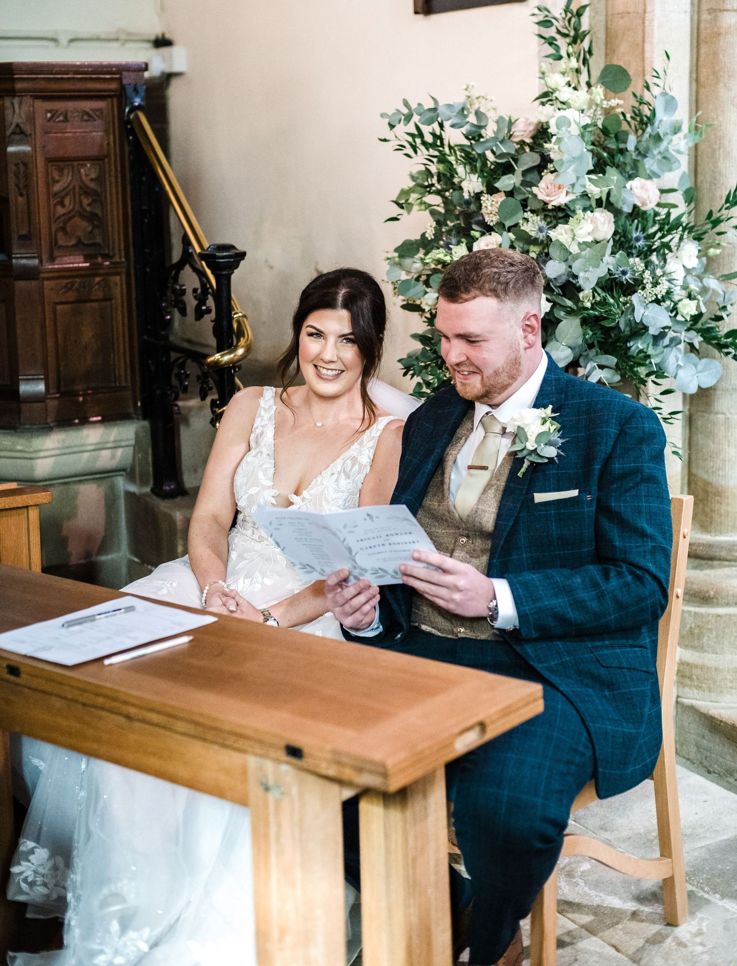 Couple signing wedding documents together at a wooden desk decorated with white floral arrangements.