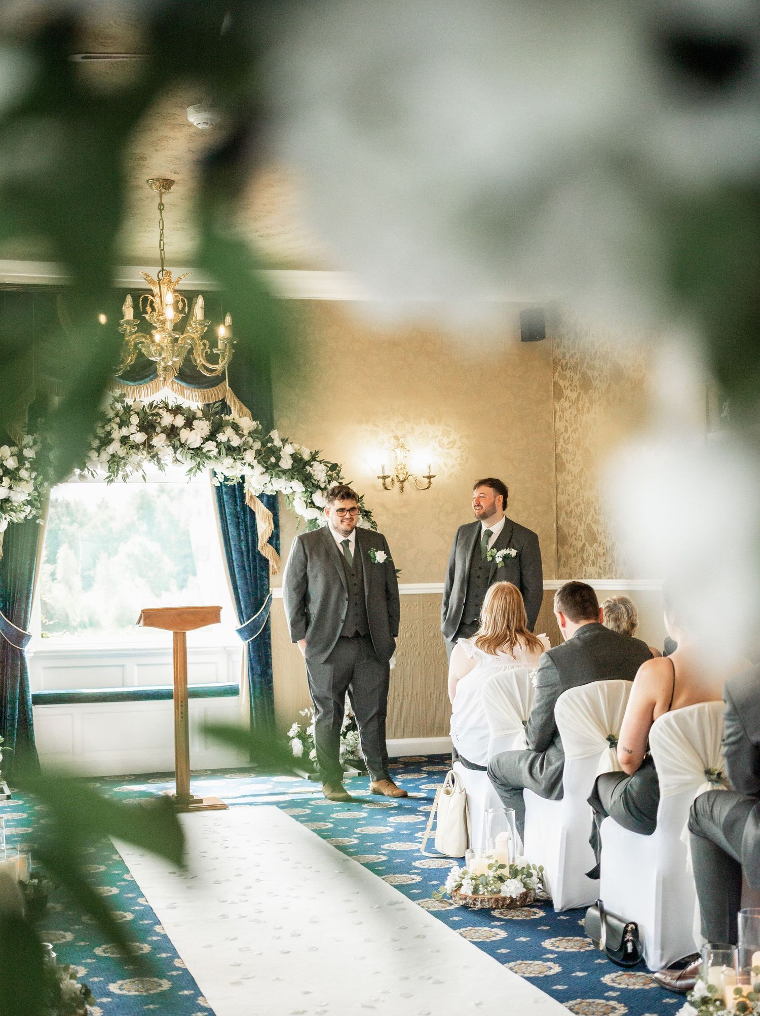 Wedding ceremony setup in elegant room with chandelier and white chairs along blue carpeted aisle.