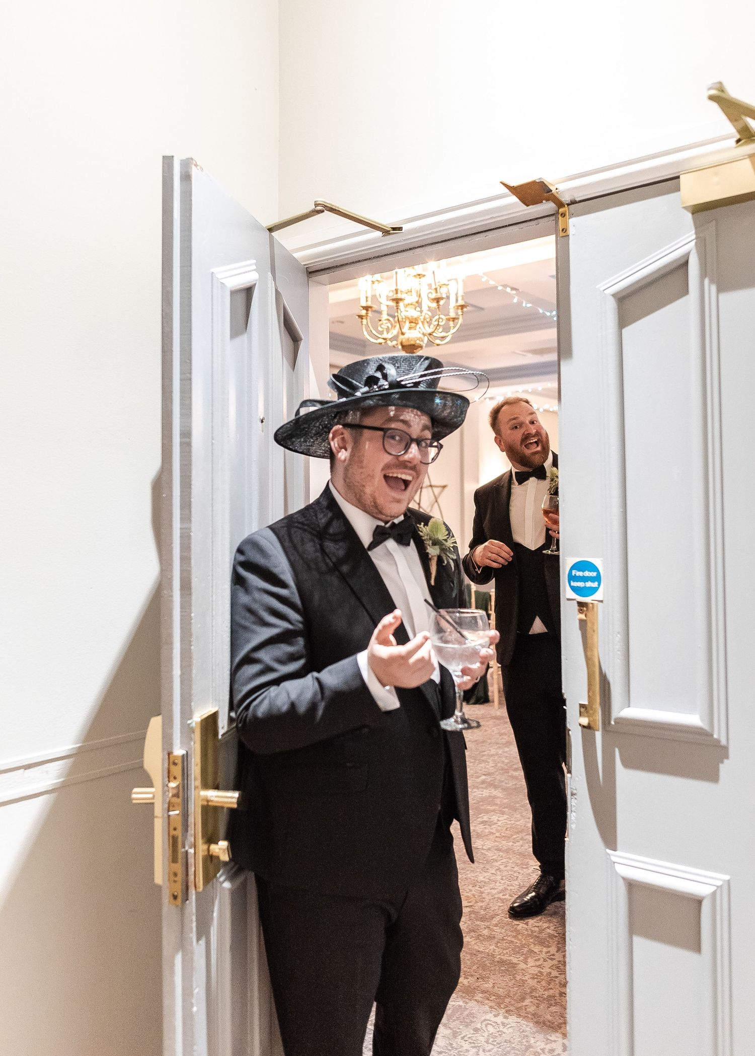 Formal event attendee wearing a decorative hat and bowtie smiles while holding a drink in an elegant hallway.