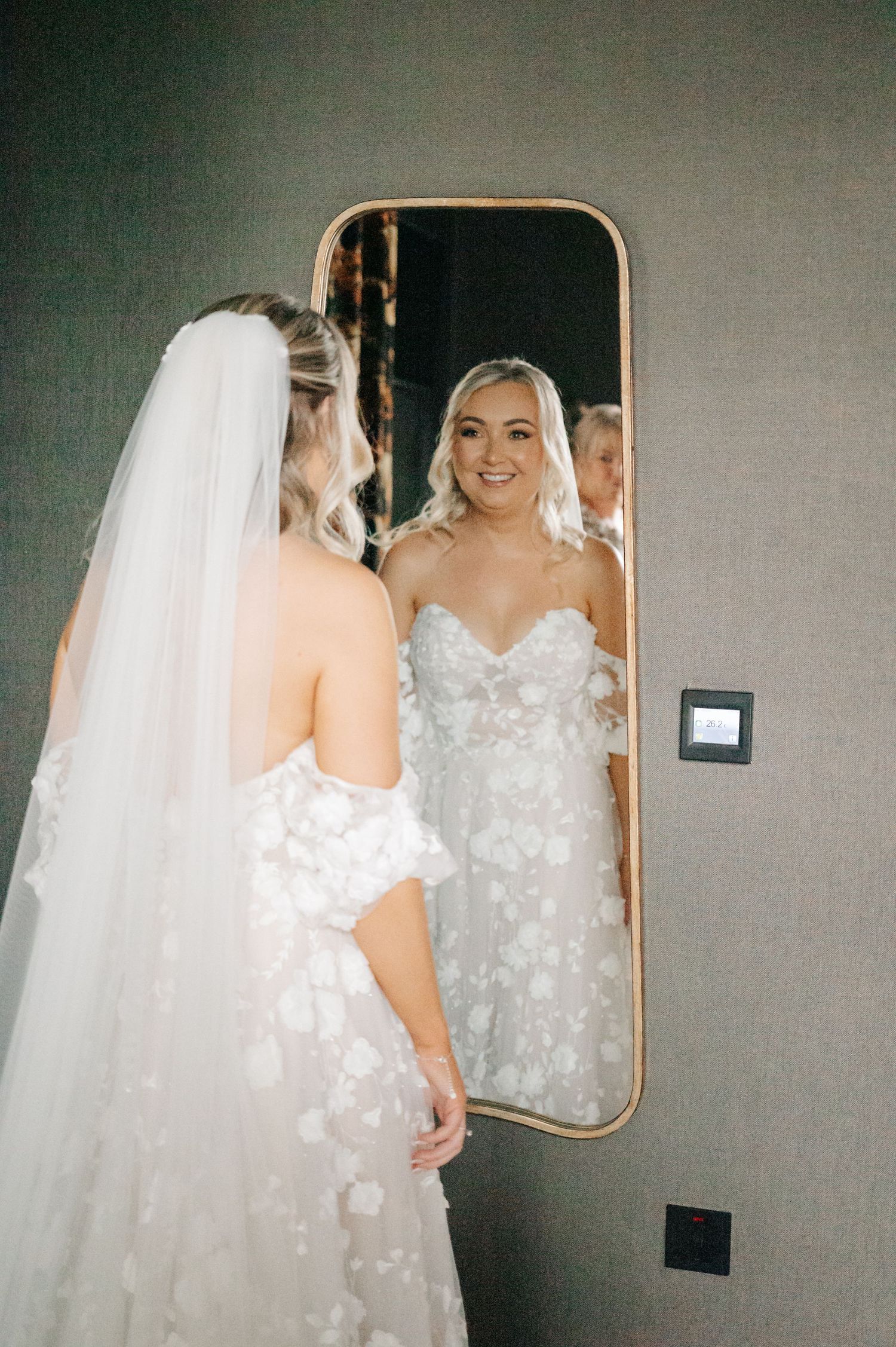 A bride wearing a delicate floral lace wedding gown admires her reflection in a mirror.