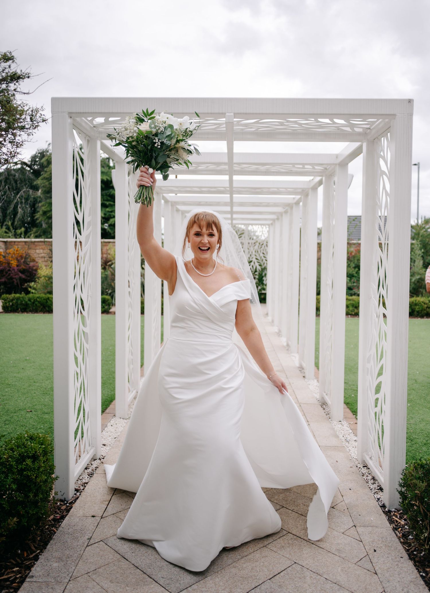 A person in a white wedding dress twirls and poses under a white pergola structure in a garden setting.