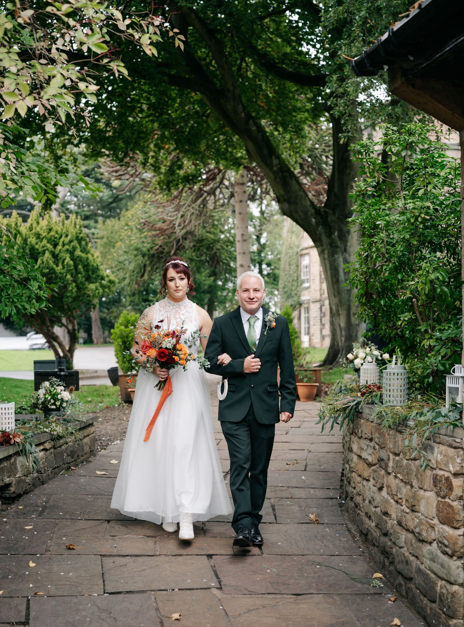 A wedding processional along a stone pathway under trees with autumn leaves.