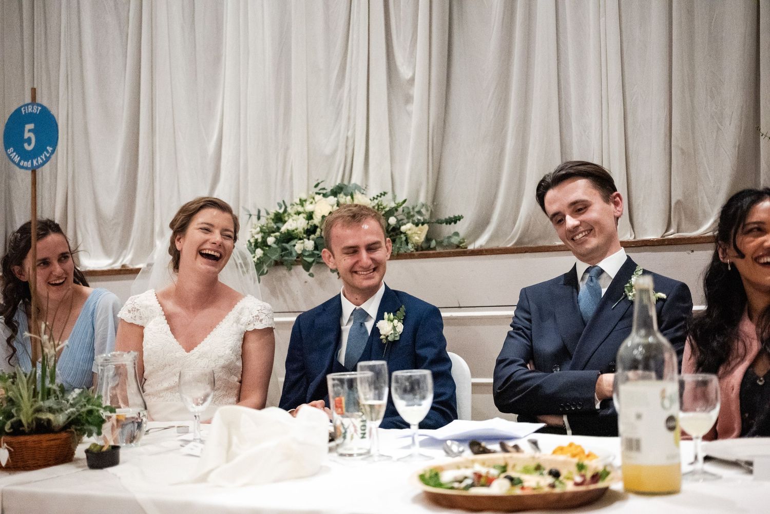 Wedding reception guests seated at long tables with white linens enjoy conversation and laughter.