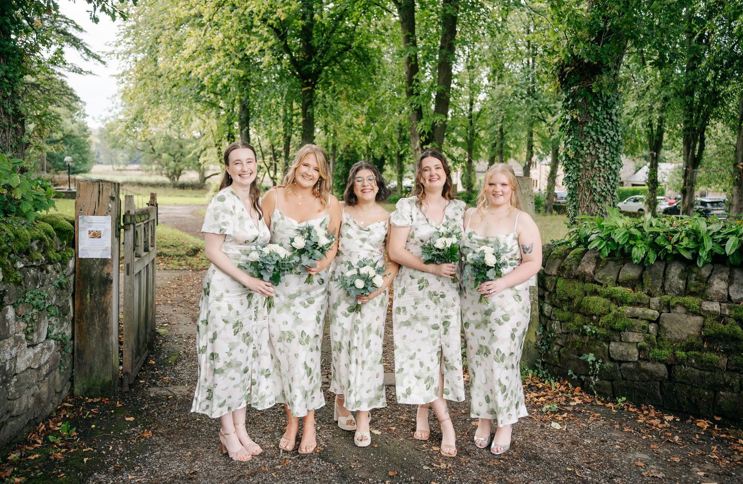 Five bridesmaids in floral dresses pose together on a scenic wooded path during an autumn wedding.