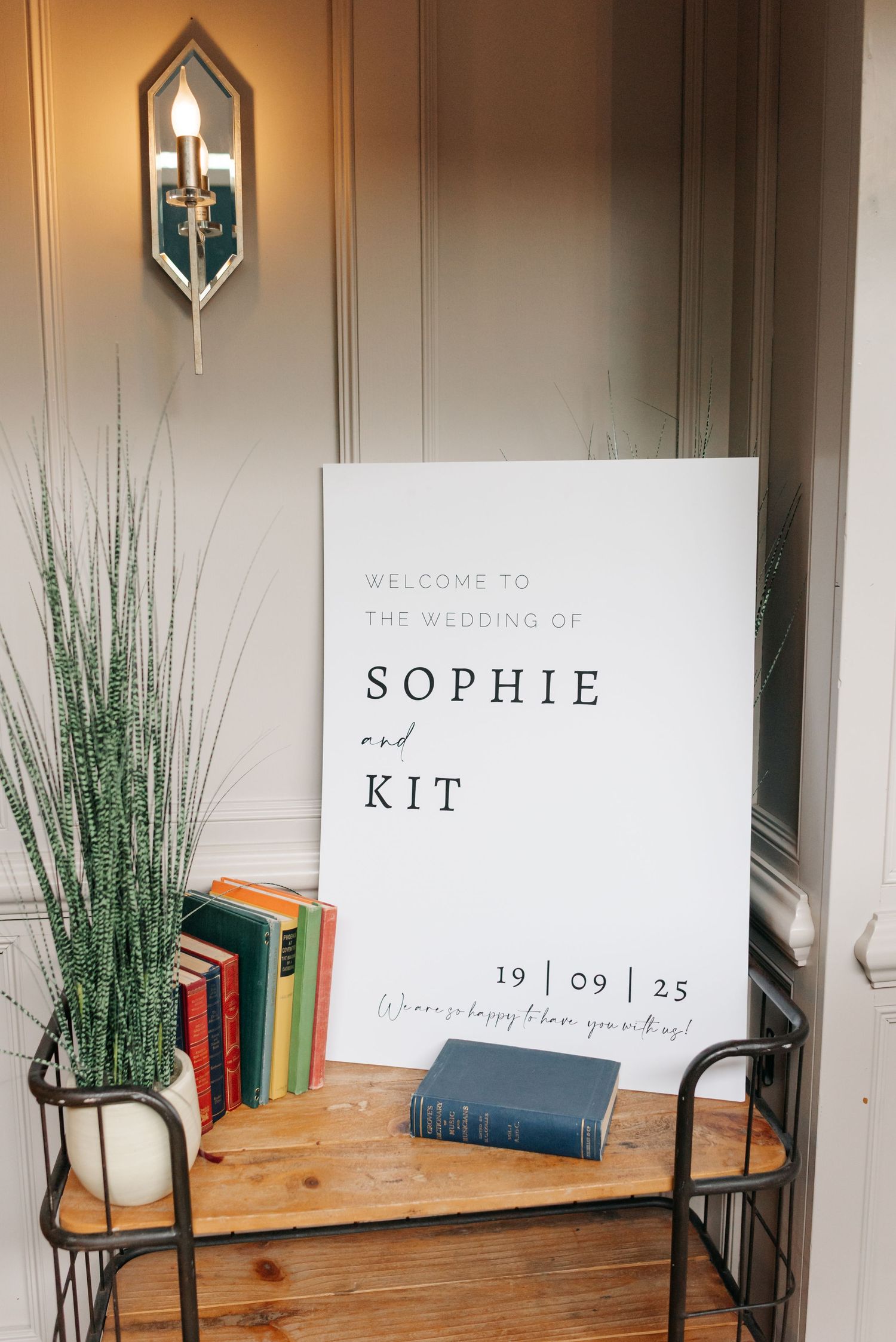 Modern wedding welcome sign display with potted plants and books on wooden shelf.