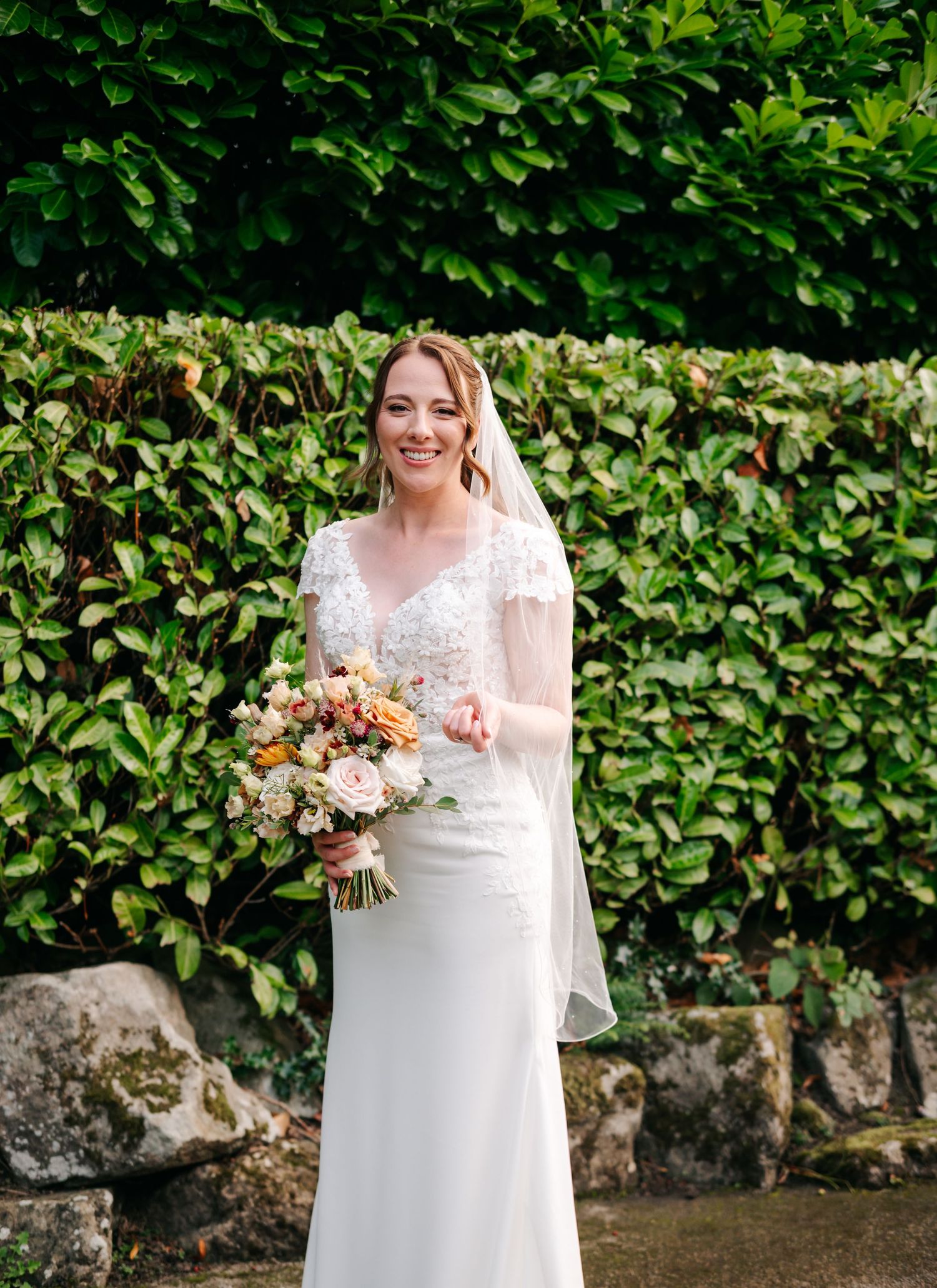 Smiling bride in an elegant white lace gown and veil holds a colorful bouquet in front of a lush green hedge and mossy stone wall.