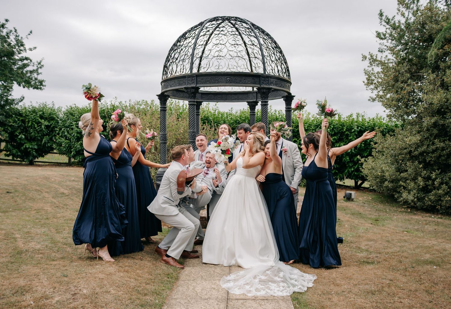 Wedding party celebrating with bride and groom near ornate gazebo, bridesmaids in navy dresses holding bouquets.