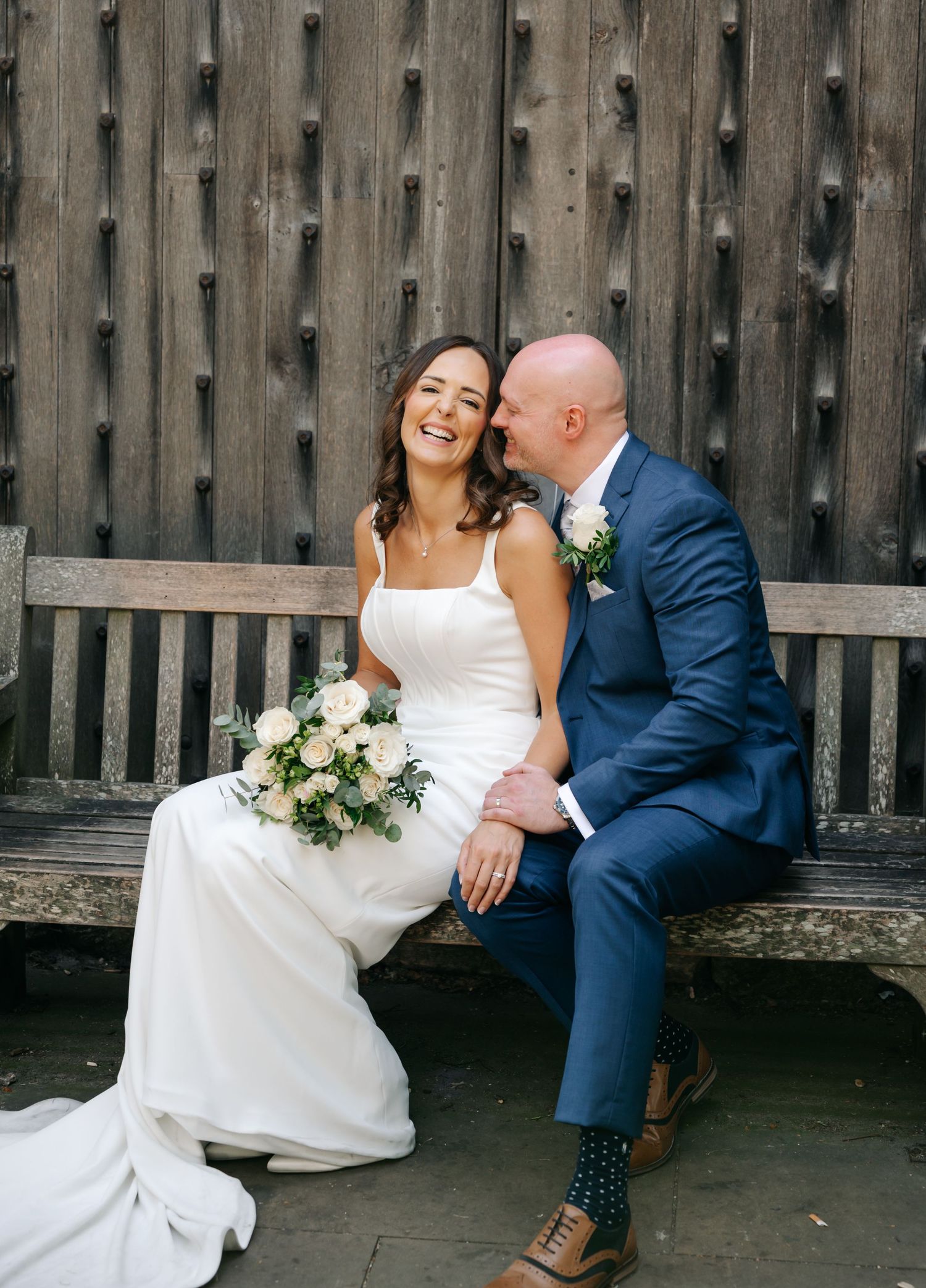 Bride in white gown holding blush bouquet sits with groom in navy suit on wooden bench, sharing a tender moment.