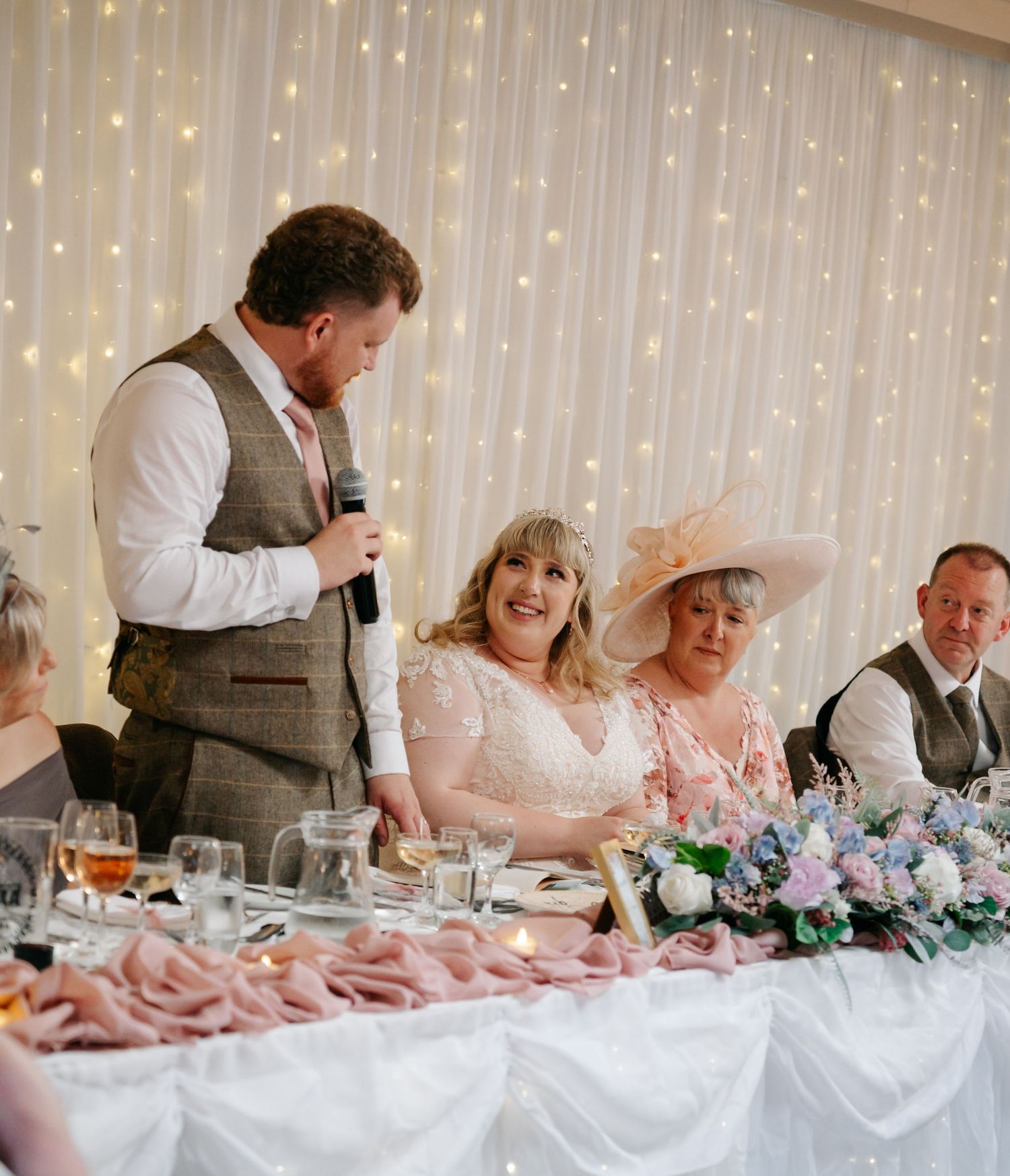 Groom giving a wedding speech at the top table, with smiling bride and guests in an elegant venue.