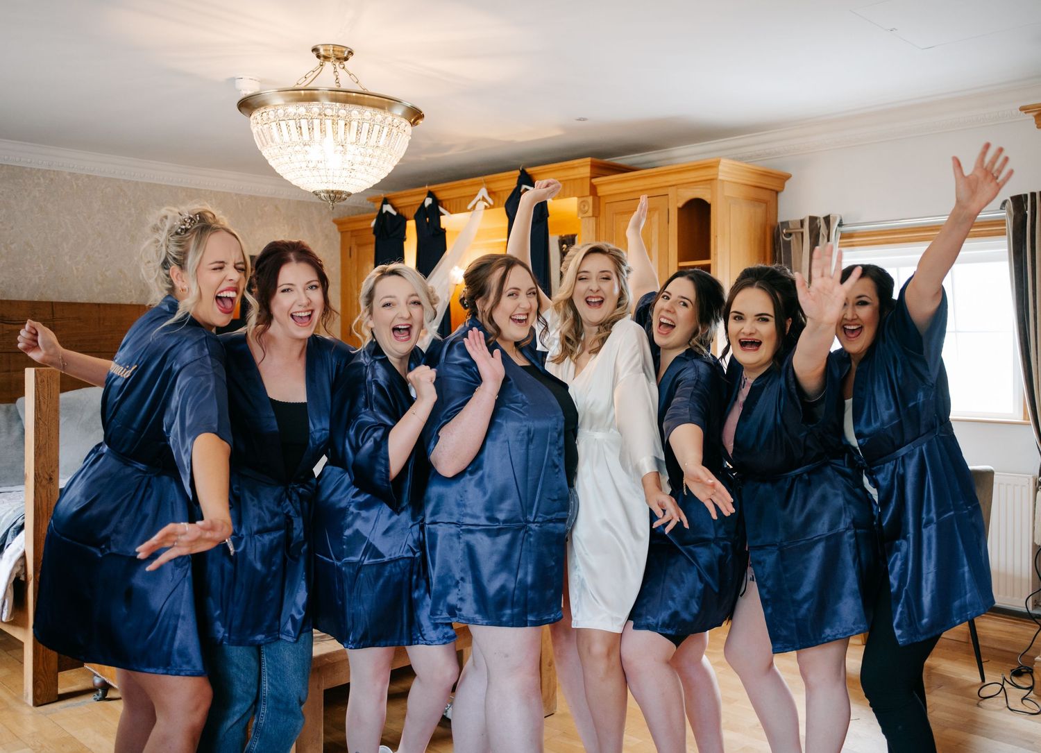 A bridal party wearing matching navy blue robes poses together for a candid group photo in a room with a chandelier.