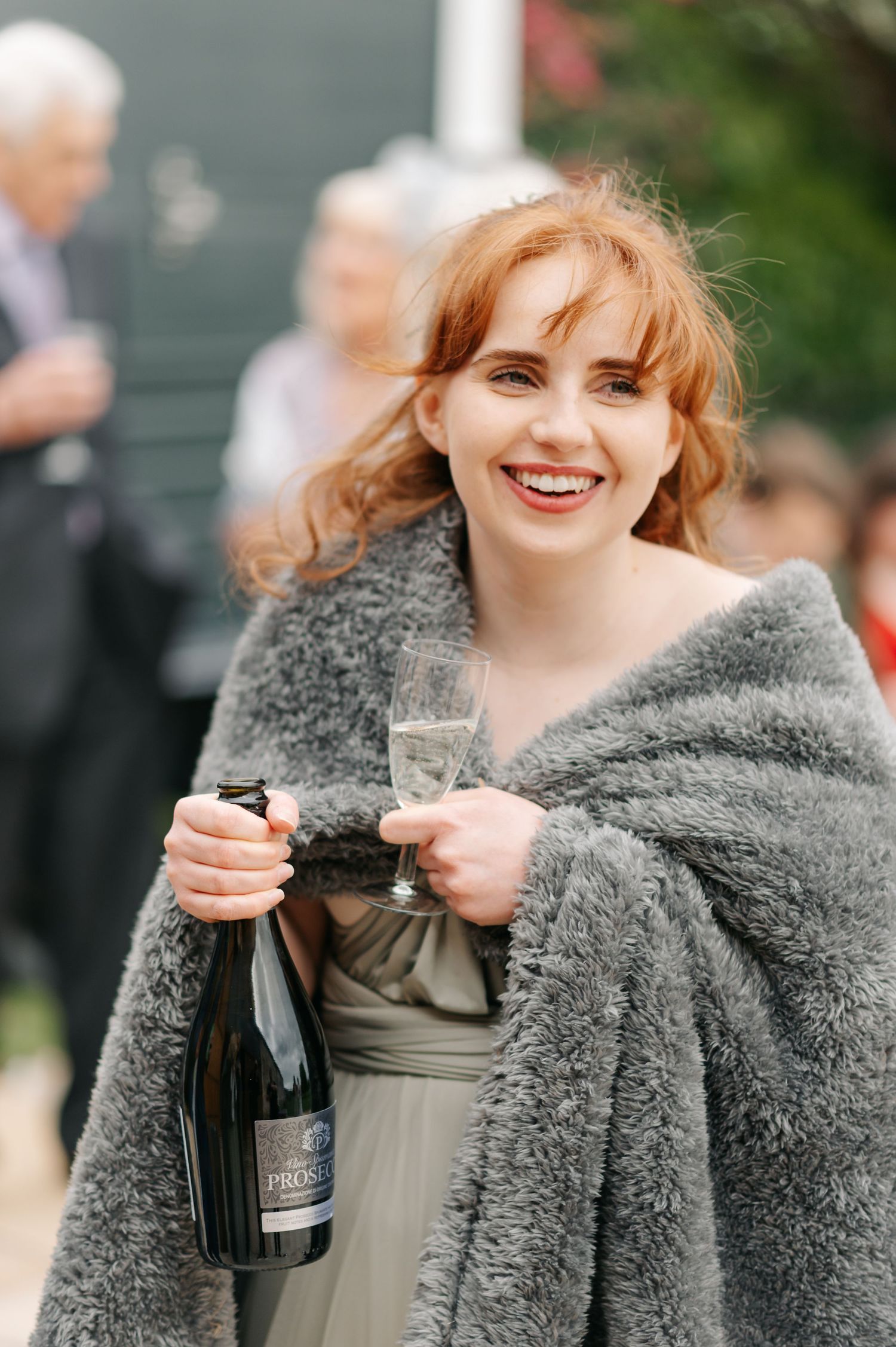 A wedding guest in a grey wrap shawl holds champagne and smiles at an outdoor reception.