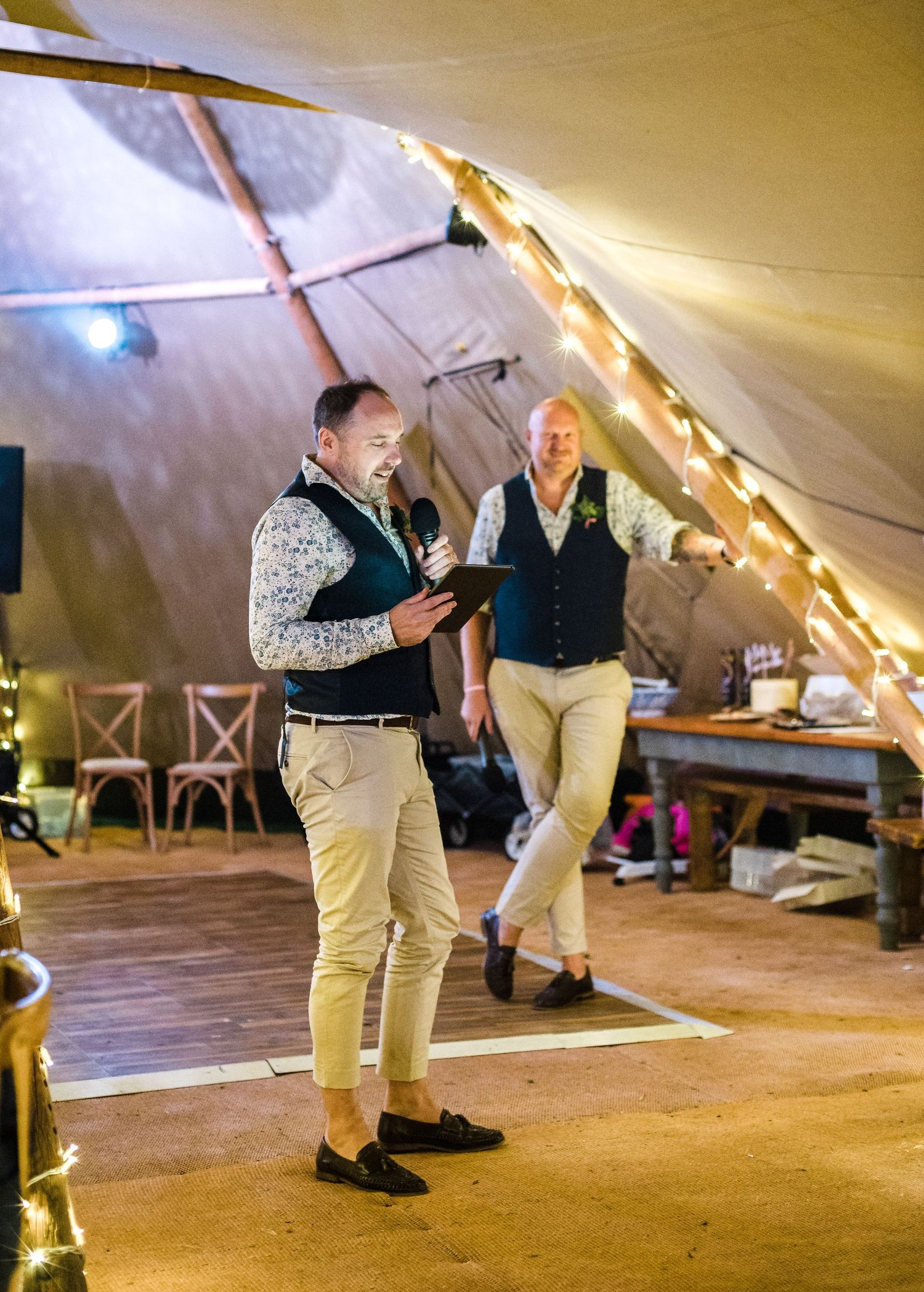 Two people in casual formal wear speaking during event inside illuminated tent venue.