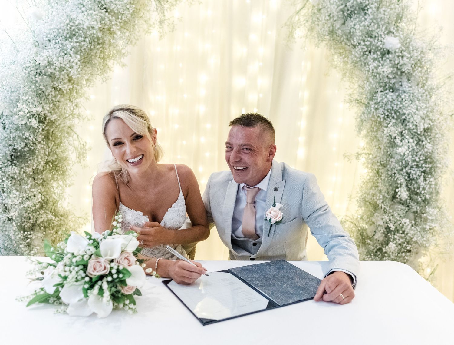 Newlyweds sign marriage license under floral arch at wedding ceremony.