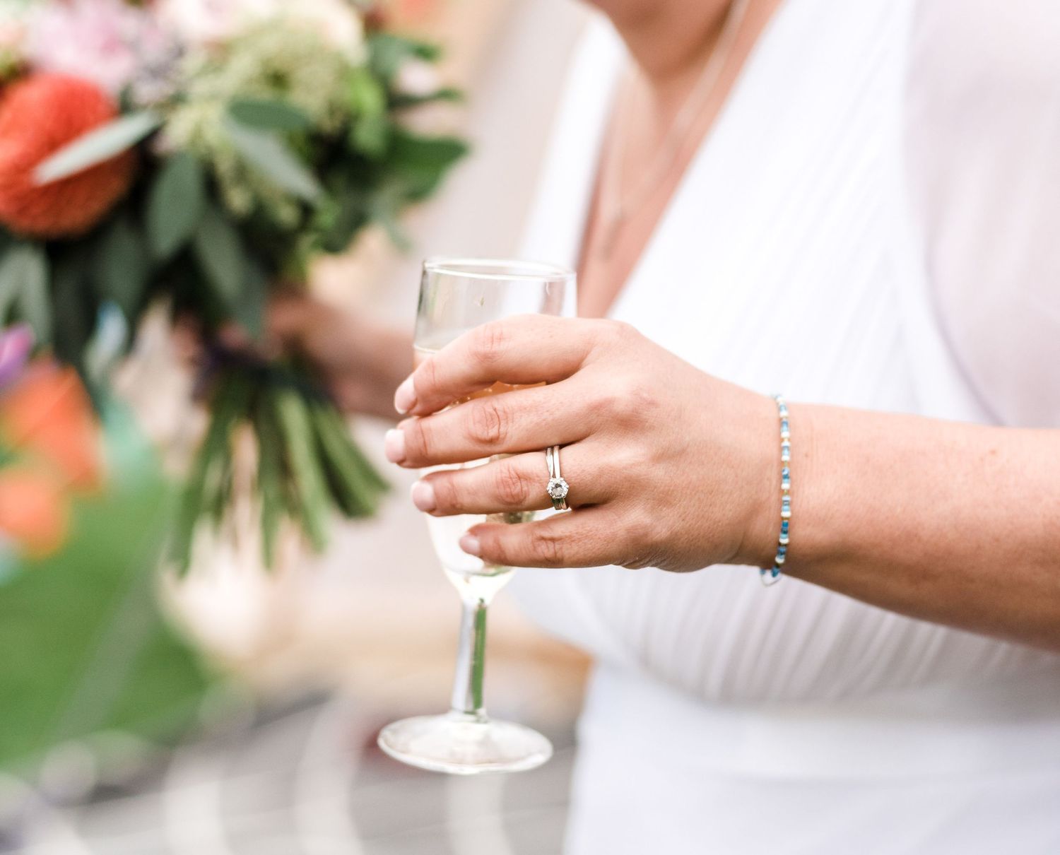 Close-up of hands holding a champagne glass with wedding ring and bouquet visible.