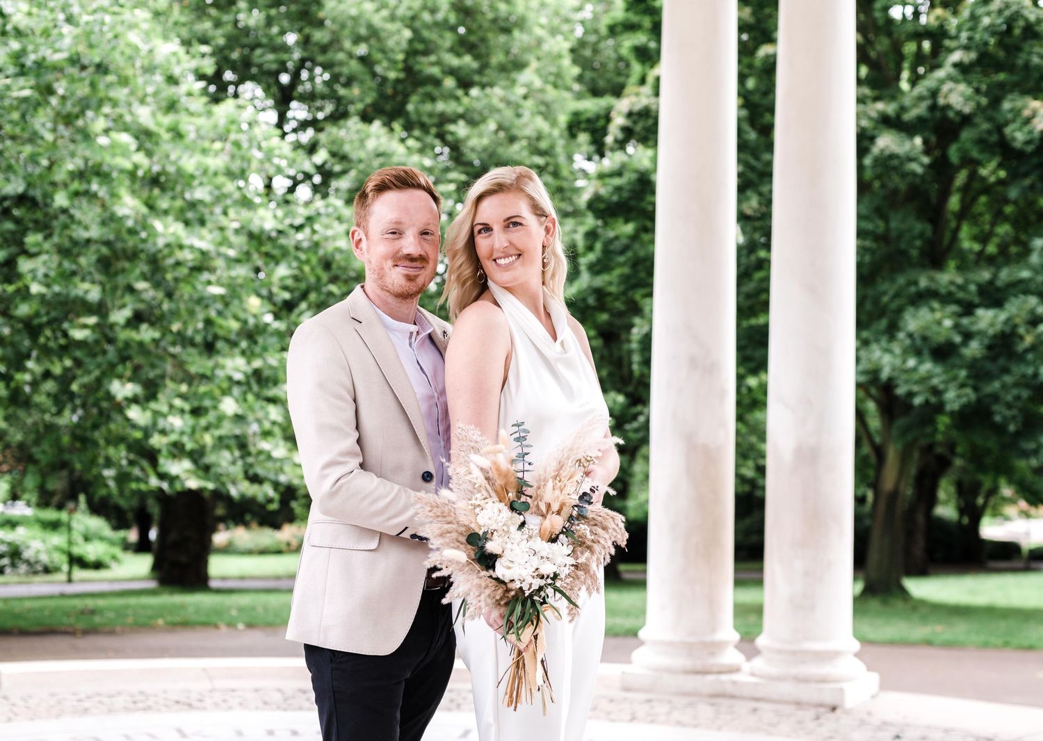 A couple poses between white columns holding a bouquet of dried flowers.