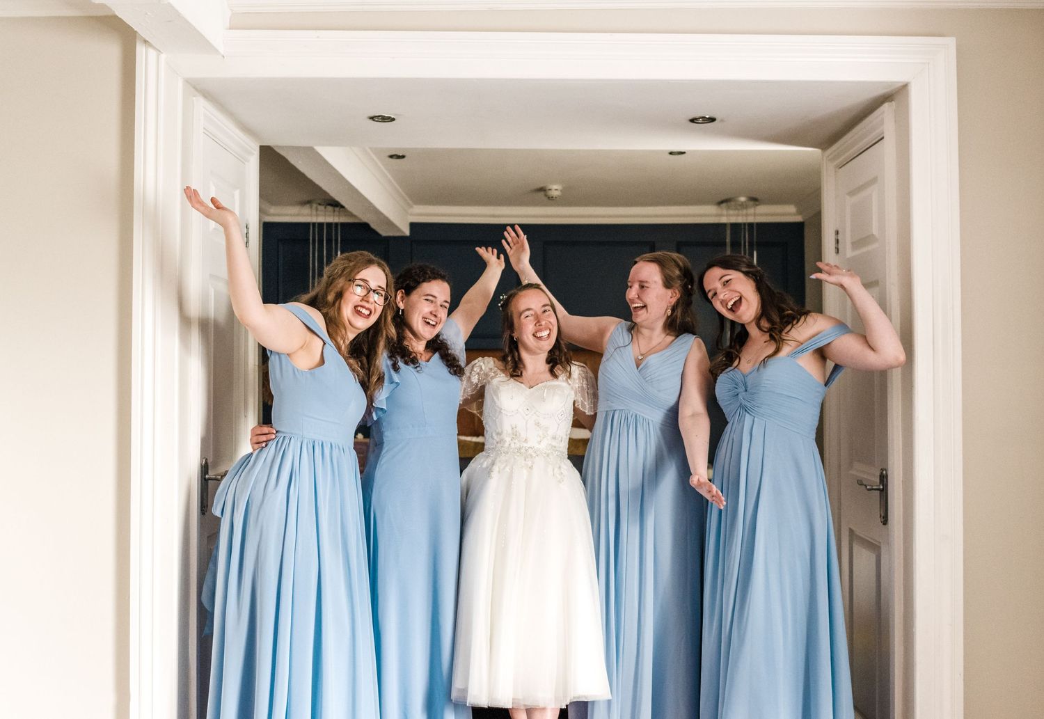 Group in matching light blue bridesmaid dresses pose excitedly with someone in white wedding dress.