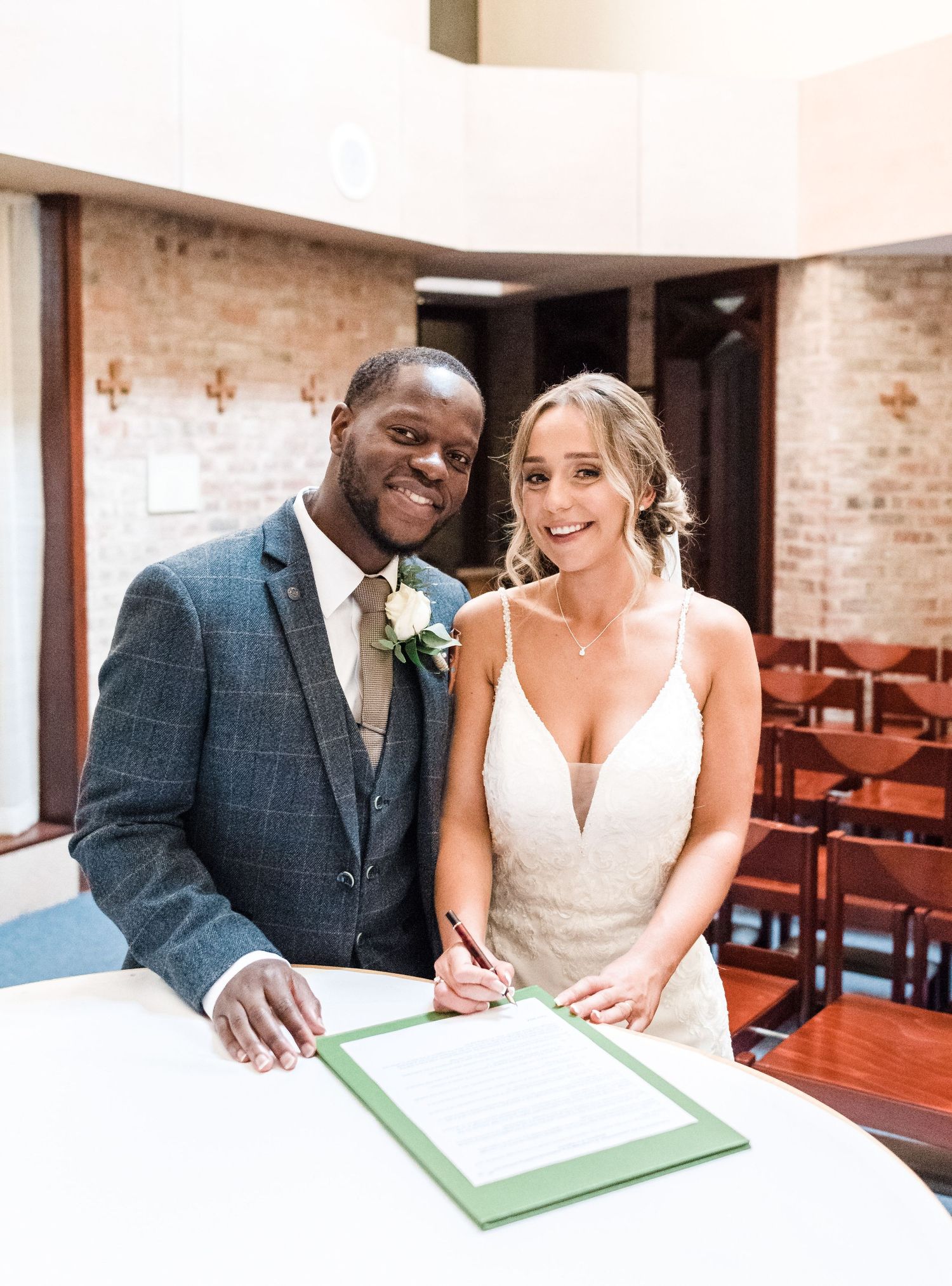 A couple smiles while signing their marriage certificate in a church with exposed brick walls.