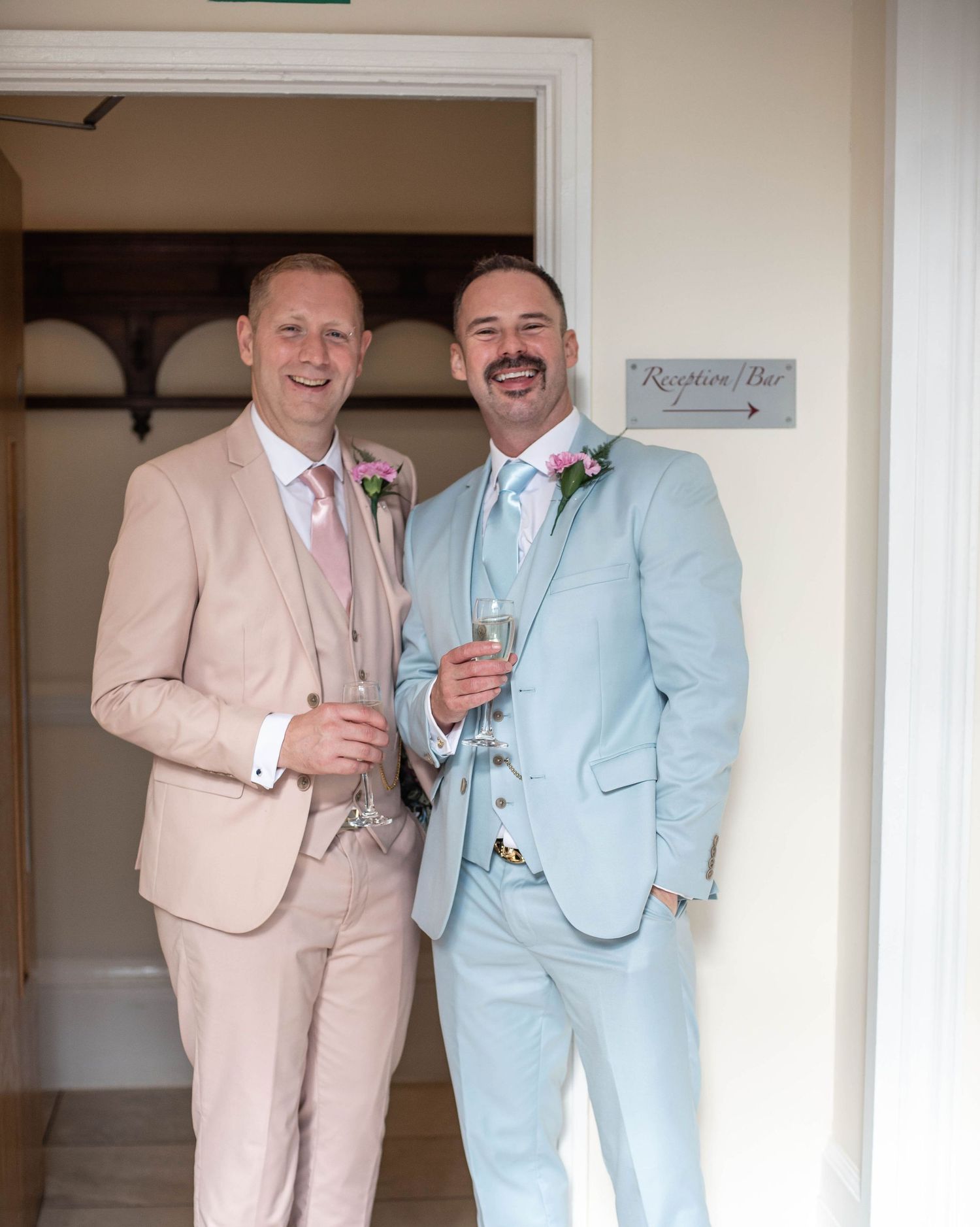 Two people in coordinating pale pink and blue suits sharing champagne.