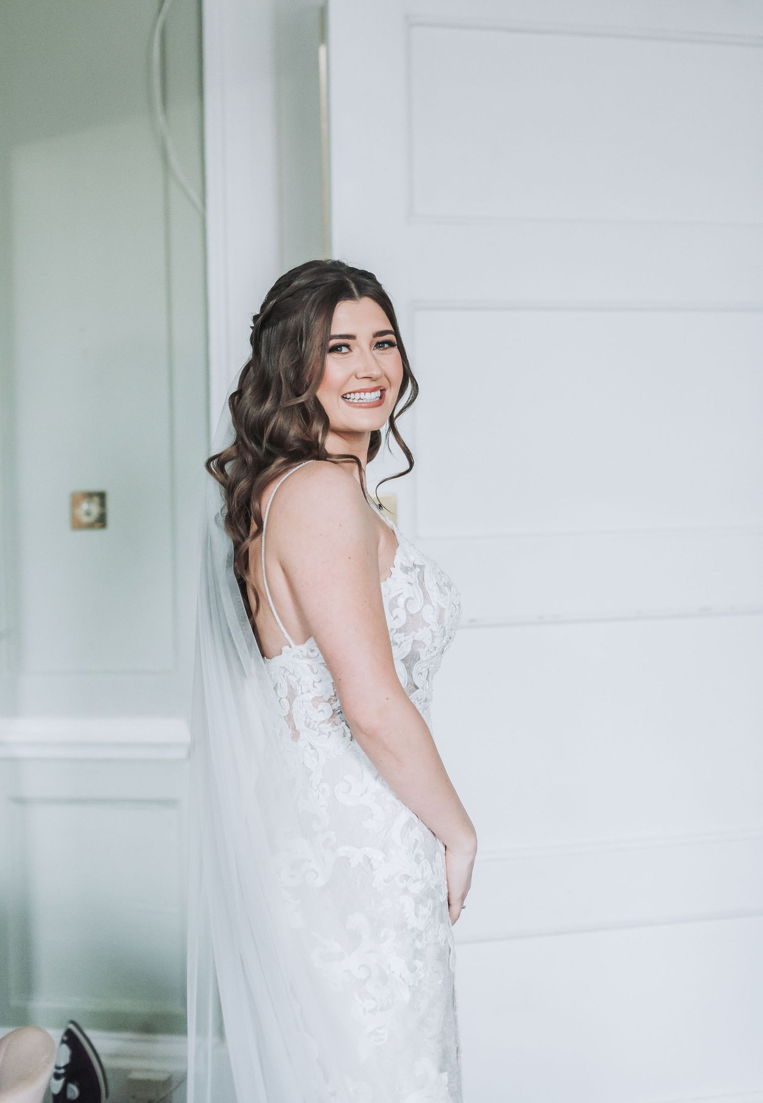 A bride in a lace wedding gown poses with a natural smile against a white wall.