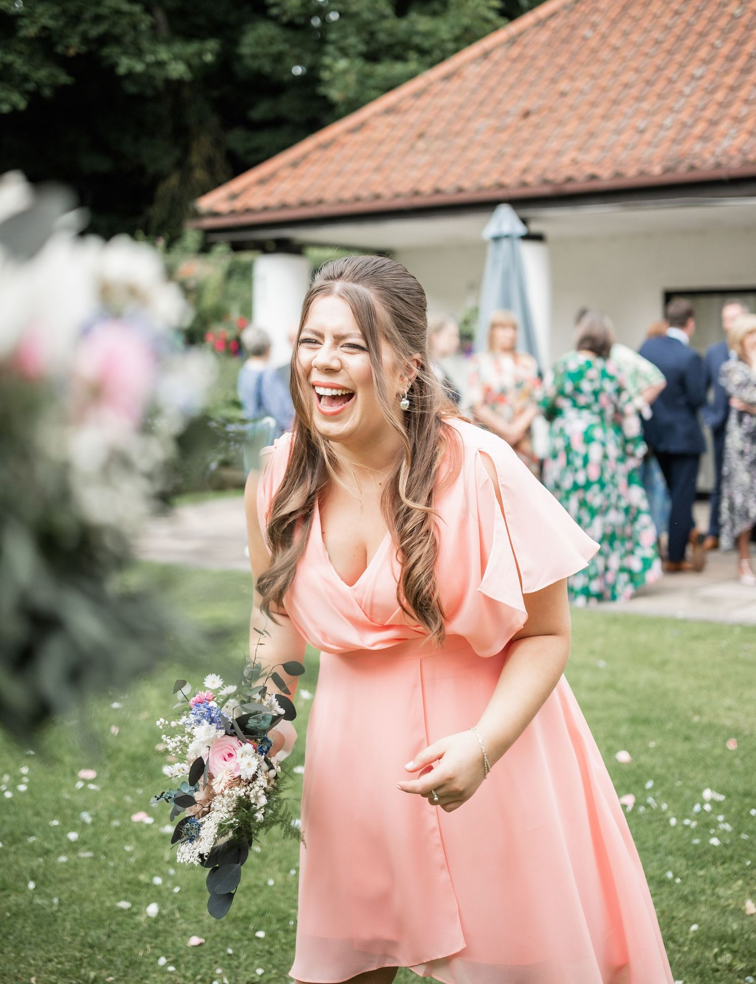 A person in a peach-colored dress laughs joyfully while holding a bouquet at an outdoor wedding celebration.