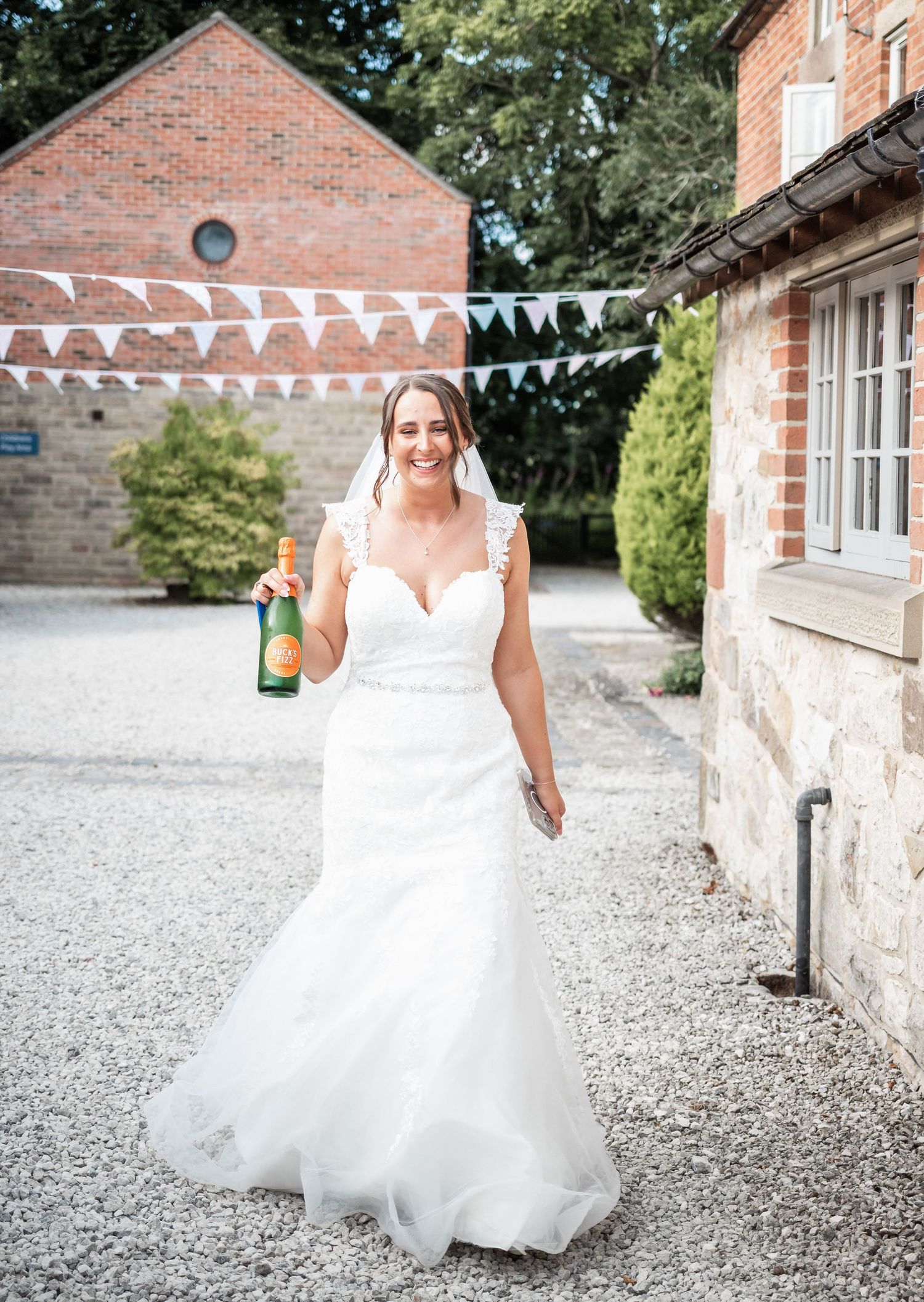 Bride in white gown holding champagne bottle on gravel path with bunting decorations.