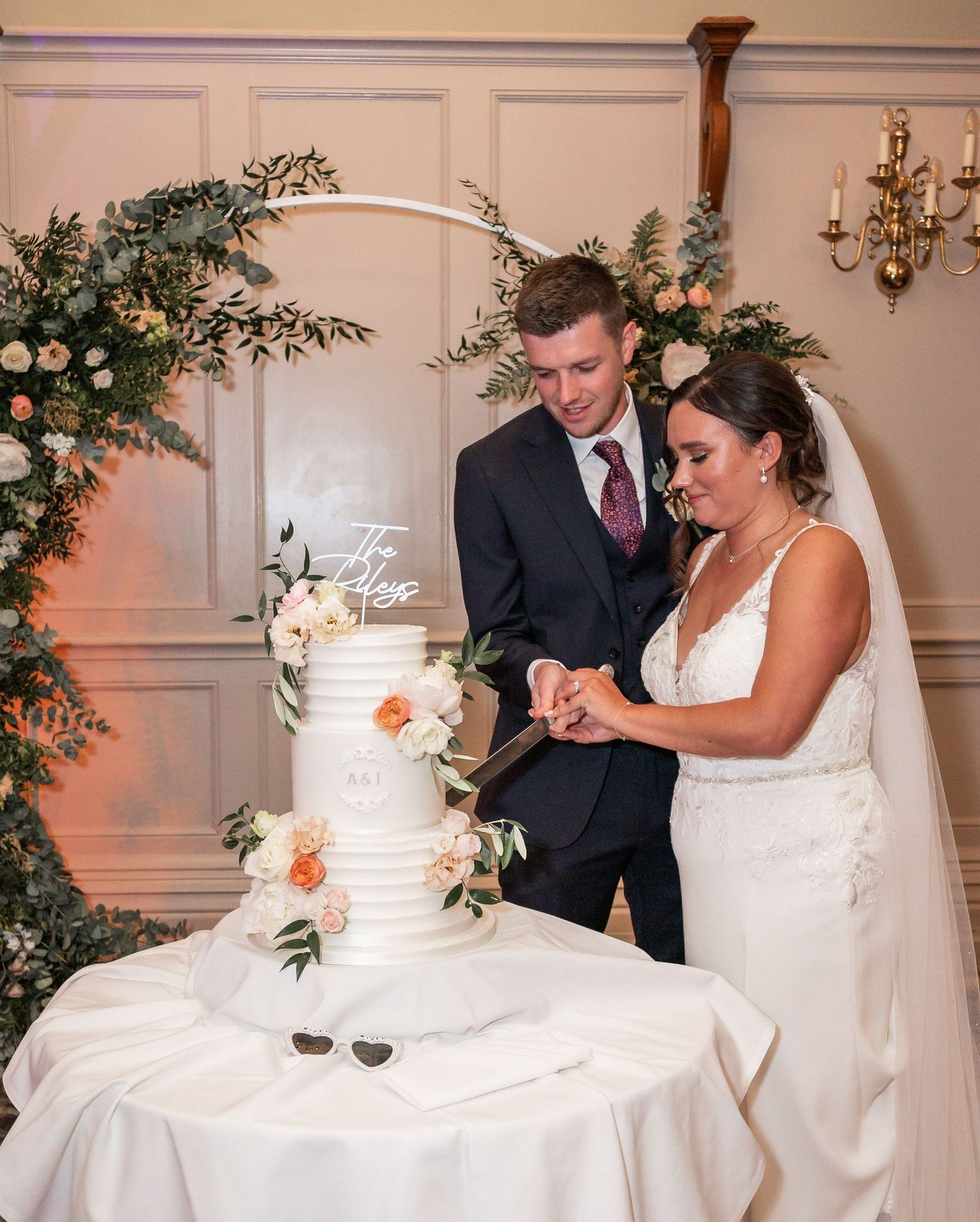 Couple cuts their elegant white wedding cake decorated with fresh flowers and greenery.