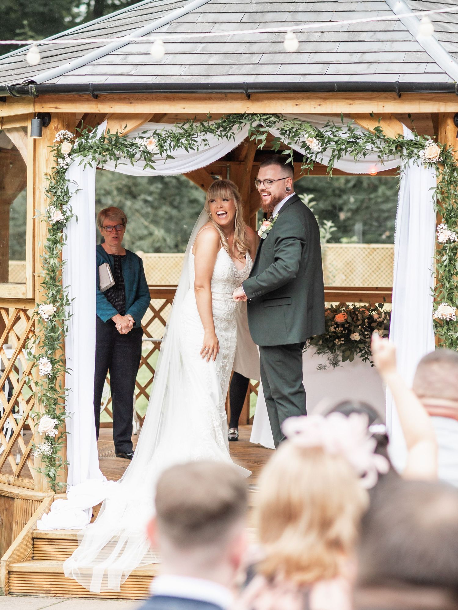 Wedding ceremony in progress at outdoor gazebo decorated with greenery and white fabric.