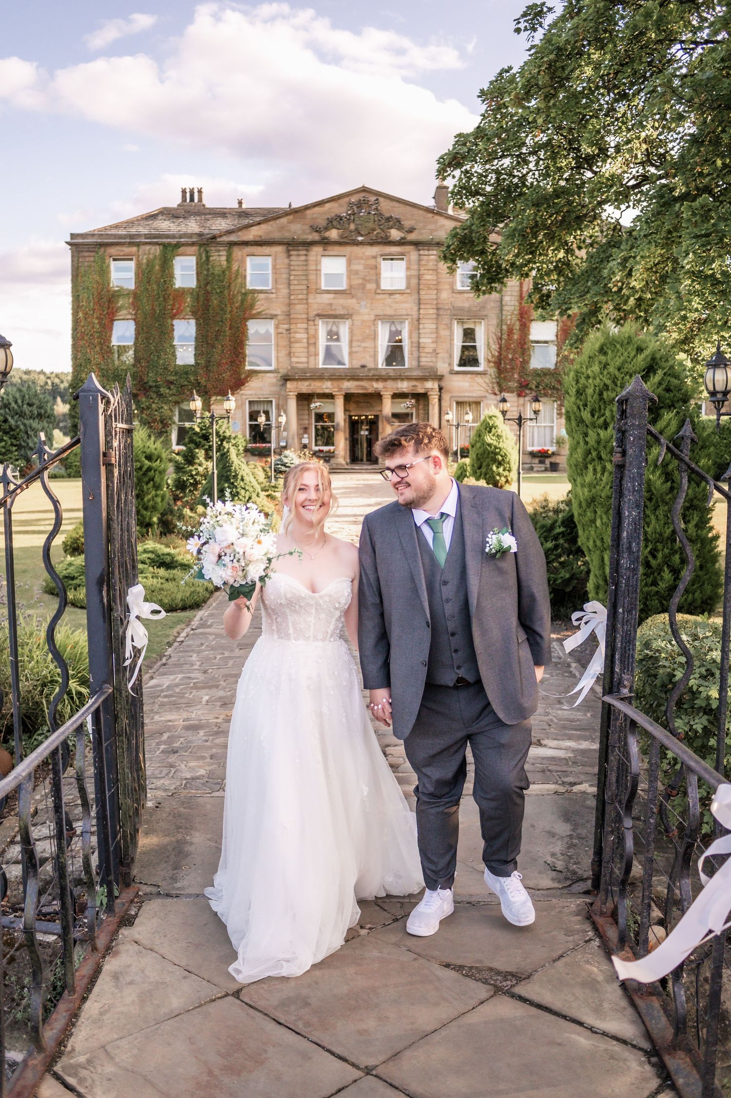 Elegant couple poses on bridge path leading to grand historic mansion.