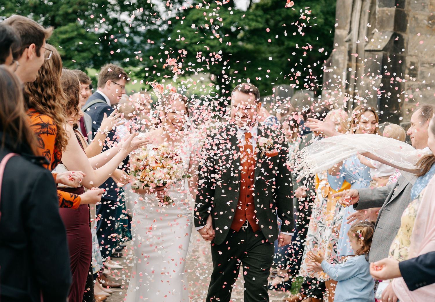 Bride and groom walk hand in hand through a crowd of guests throwing colorful confetti at an outdoor wedding celebration.
