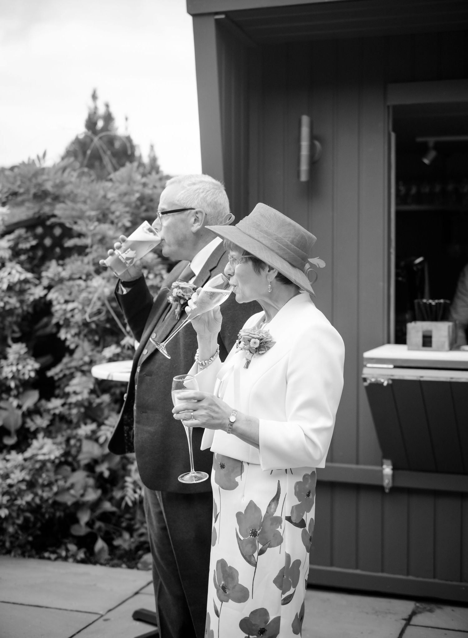 Elegant older couple in formal attire raising champagne glasses in a toast outdoors, the woman wearing a hat and floral skirt.