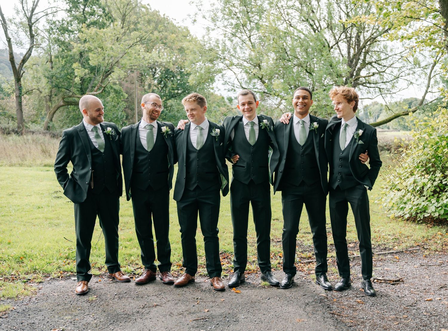 A group of groomsmen in matching black suits and green ties pose together on a garden path.