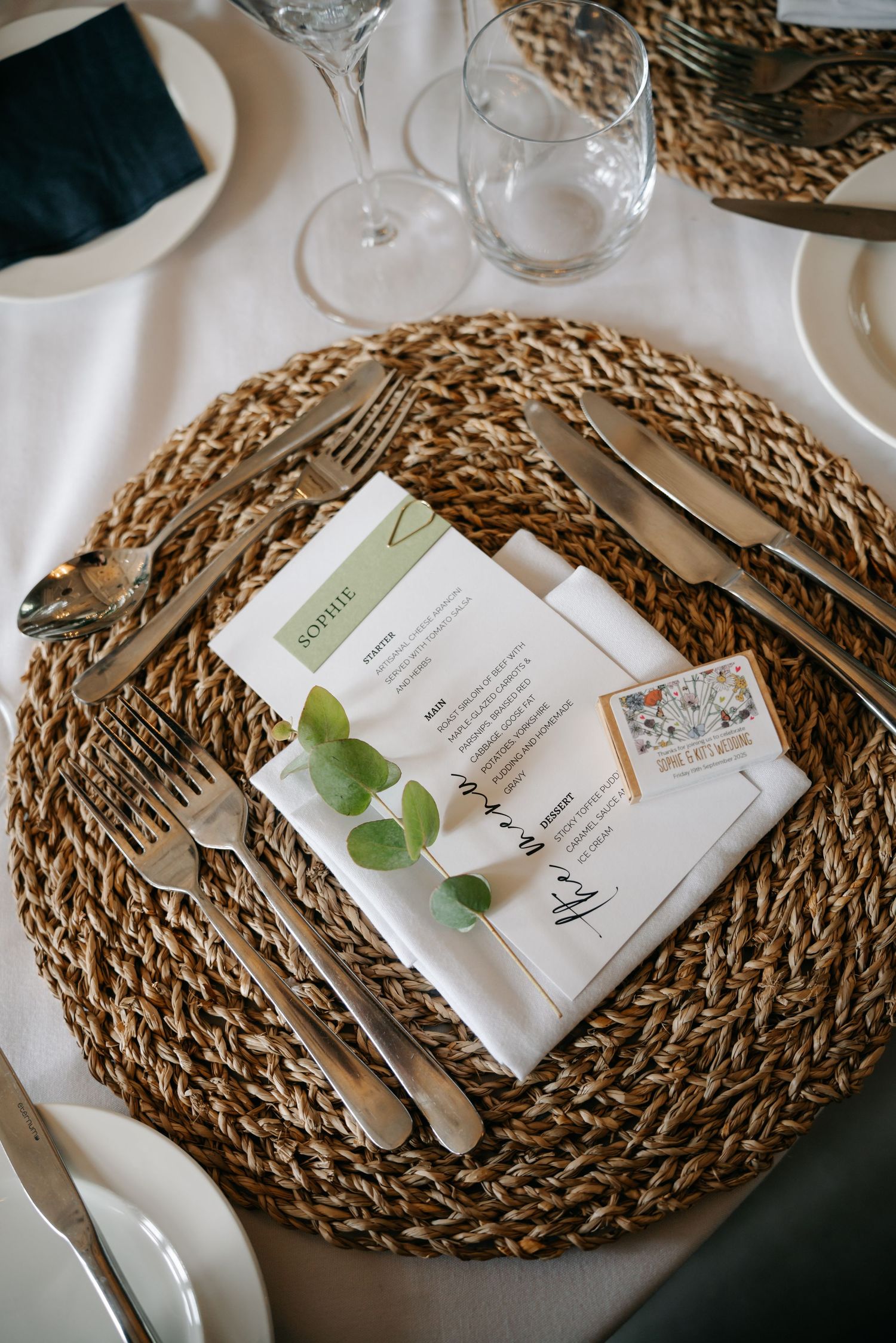 Elegant table setting with woven placemats, menu cards and eucalyptus leaves on white tablecloth.