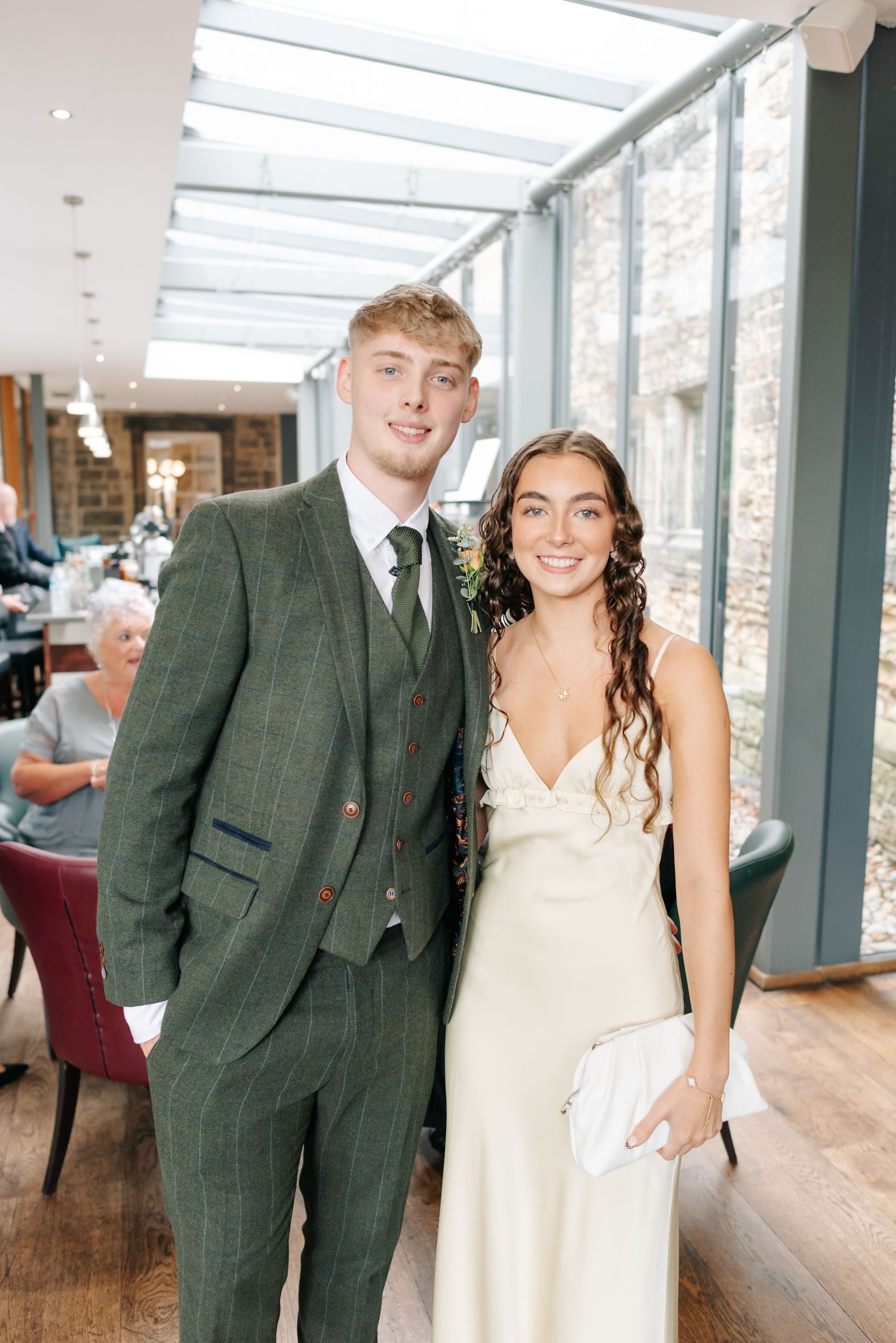 Wedding couple poses in front of large windows in a bright indoor venue wearing dark suit and cream dress.