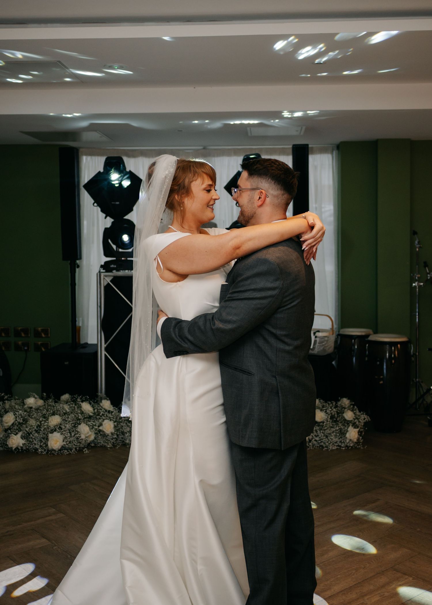 Newlyweds share their first dance on a wooden floor surrounded by floral decorations.