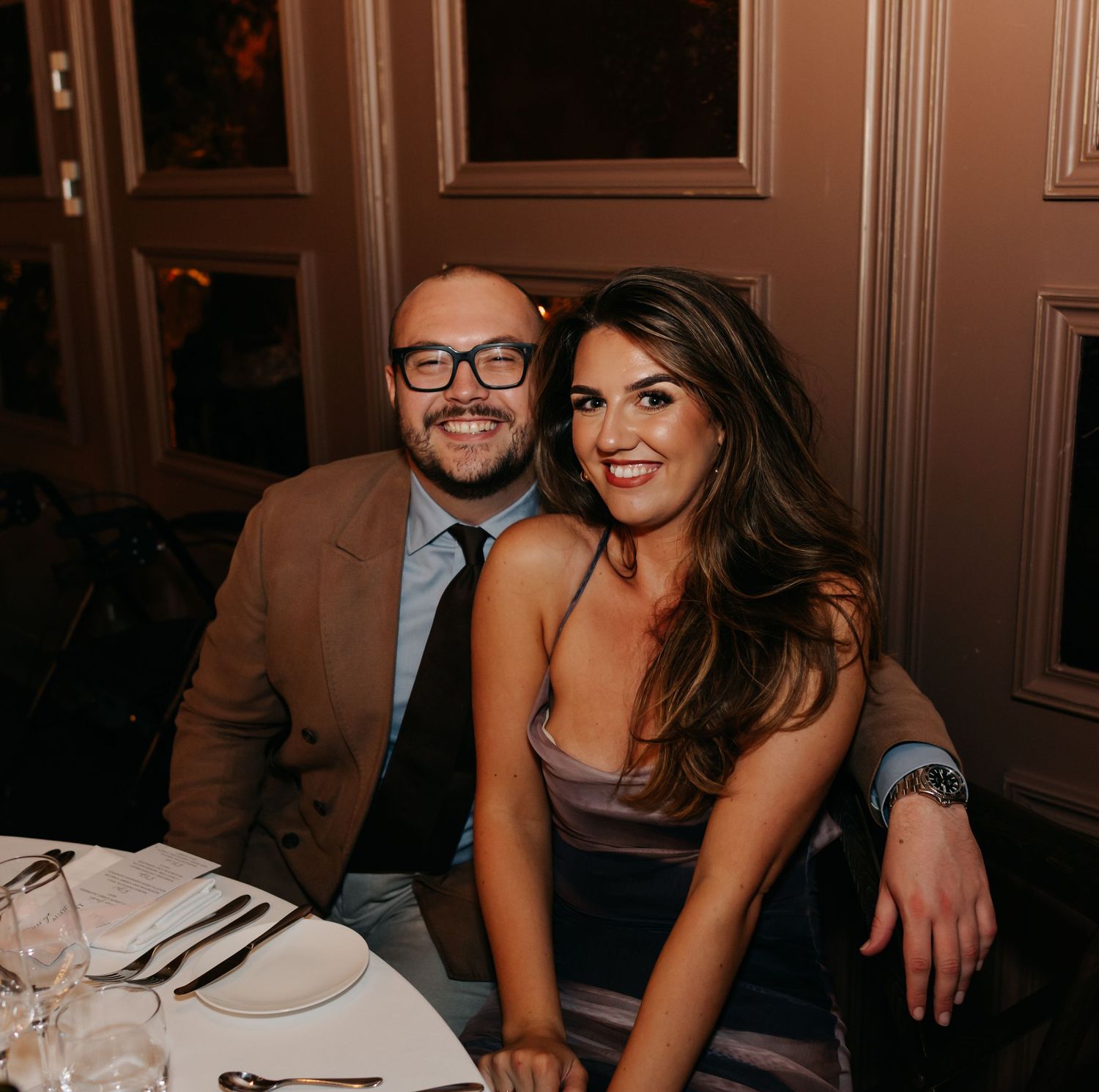 Wedding reception guests enjoying dinner at elegantly set tables in warm lighting.