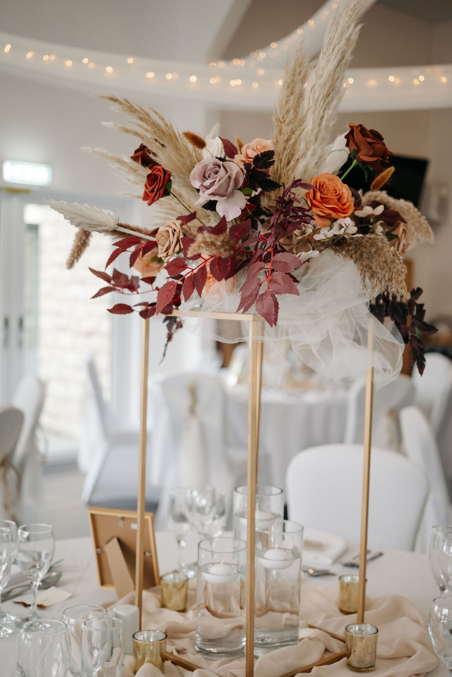 Elevated floral arrangements with dried pampas grass and autumn-colored blooms on wedding reception tables.