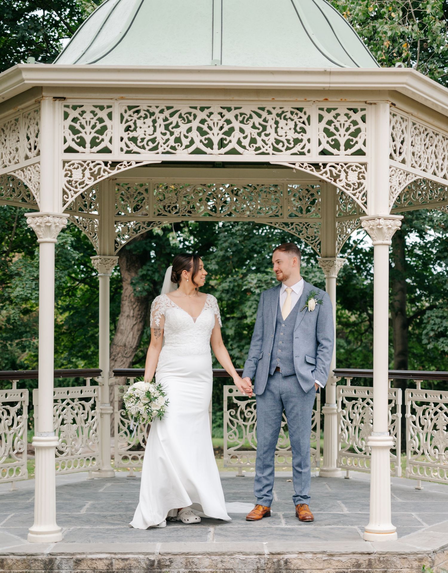 Couple poses in an ornate white Victorian-style gazebo surrounded by lush greenery.
