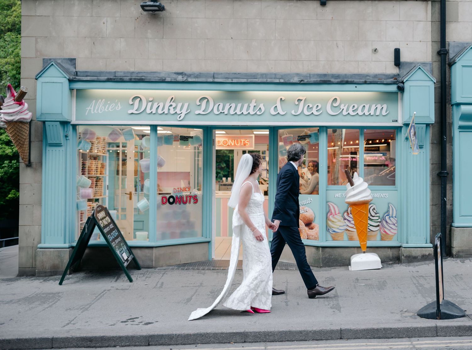 A couple walks past a row of pastel-colored storefronts including a donut and ice cream shop.